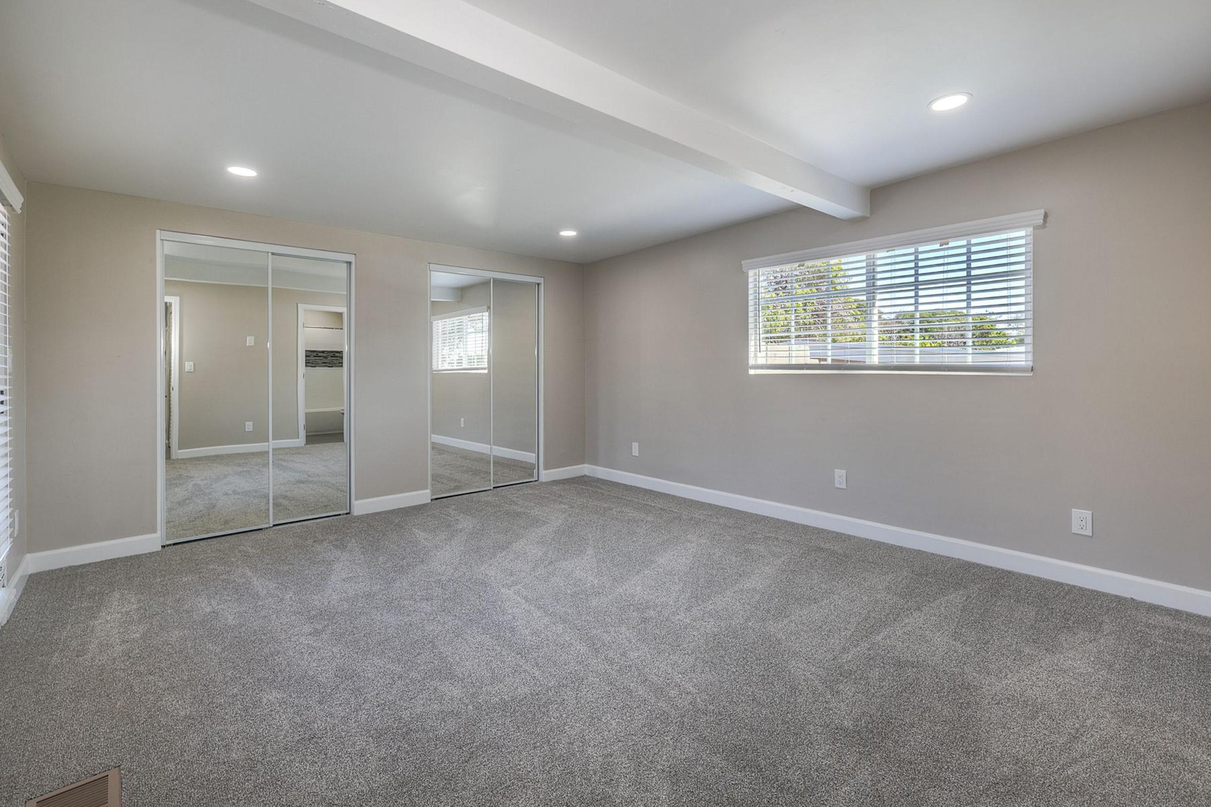 A spacious room with beige walls and carpeted flooring. It features large sliding mirrors on one wall and a window with white blinds allowing natural light. The room is empty, creating an open and airy atmosphere, ideal for various uses.