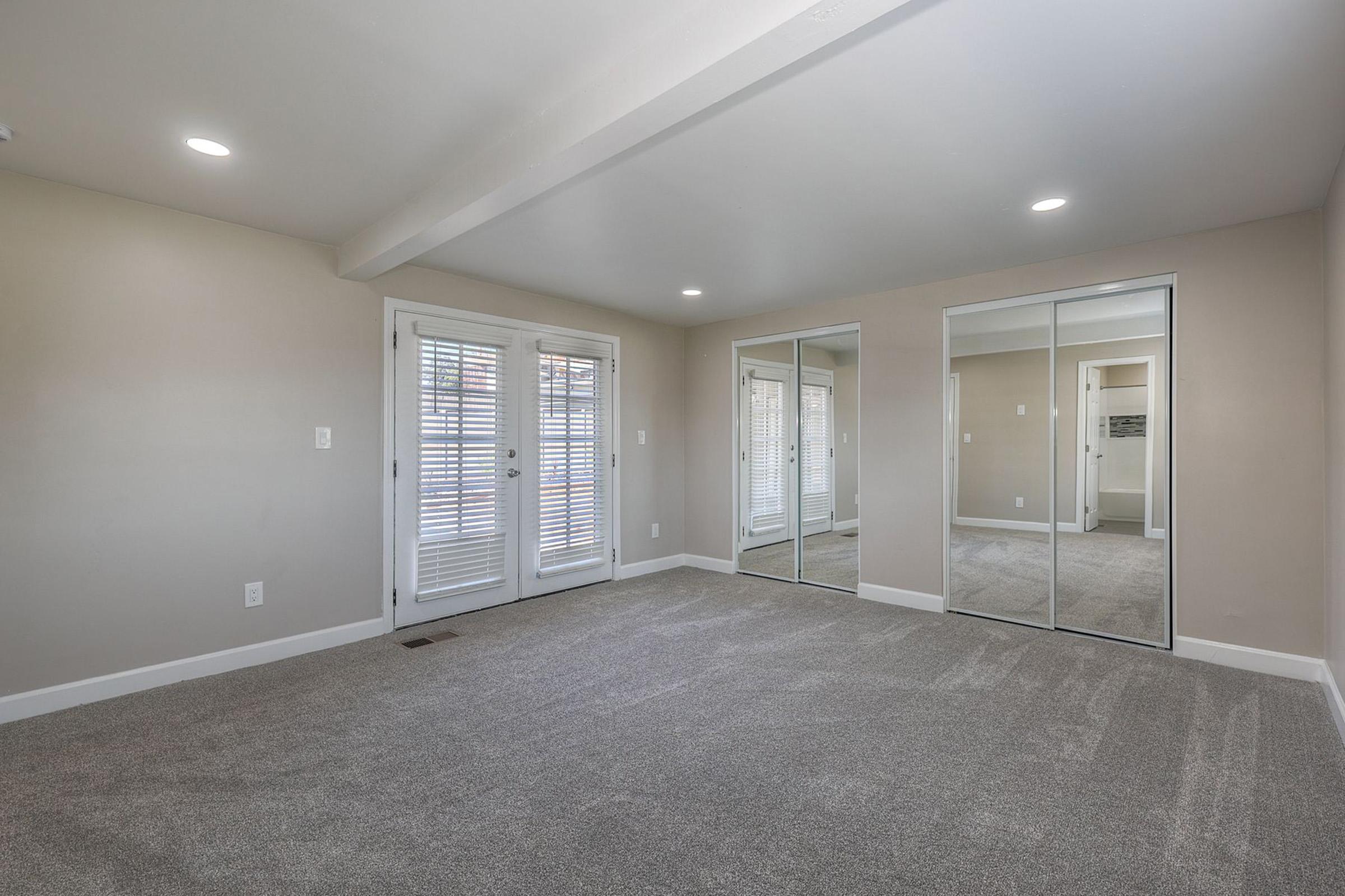 A spacious, well-lit room featuring light gray carpet, neutral walls, and sliding glass doors leading to a balcony or outdoor area. There are two large mirrored closets on one side, providing ample storage space. The ceiling has recessed lighting for a bright atmosphere.
