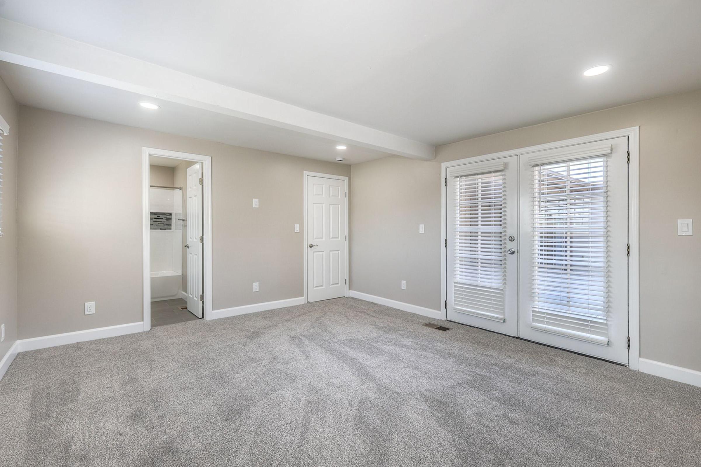 A spacious, light-filled room featuring neutral gray walls and plush carpet. Two sets of French doors with blinds open to a bright exterior, while a door on the left leads to a bathroom. The overall design is modern and minimalistic, ideal for various uses.