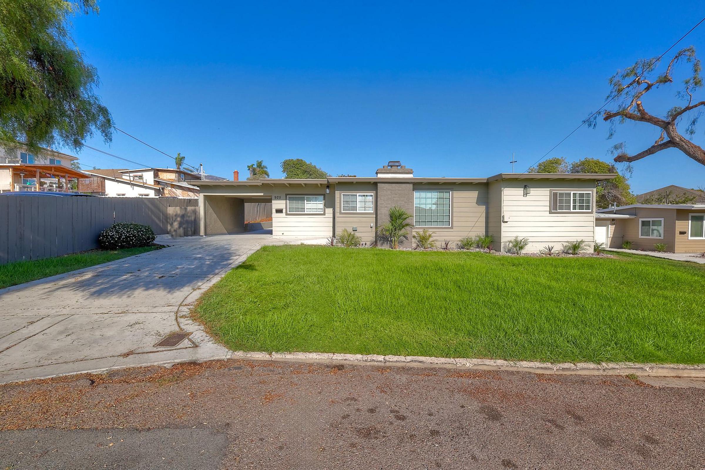 A single-story residential home with a light-colored exterior and a manicured lawn. The house features a driveway leading to a carport, surrounded by a modern landscape. In the background, there are neighboring homes and a clear blue sky overhead.