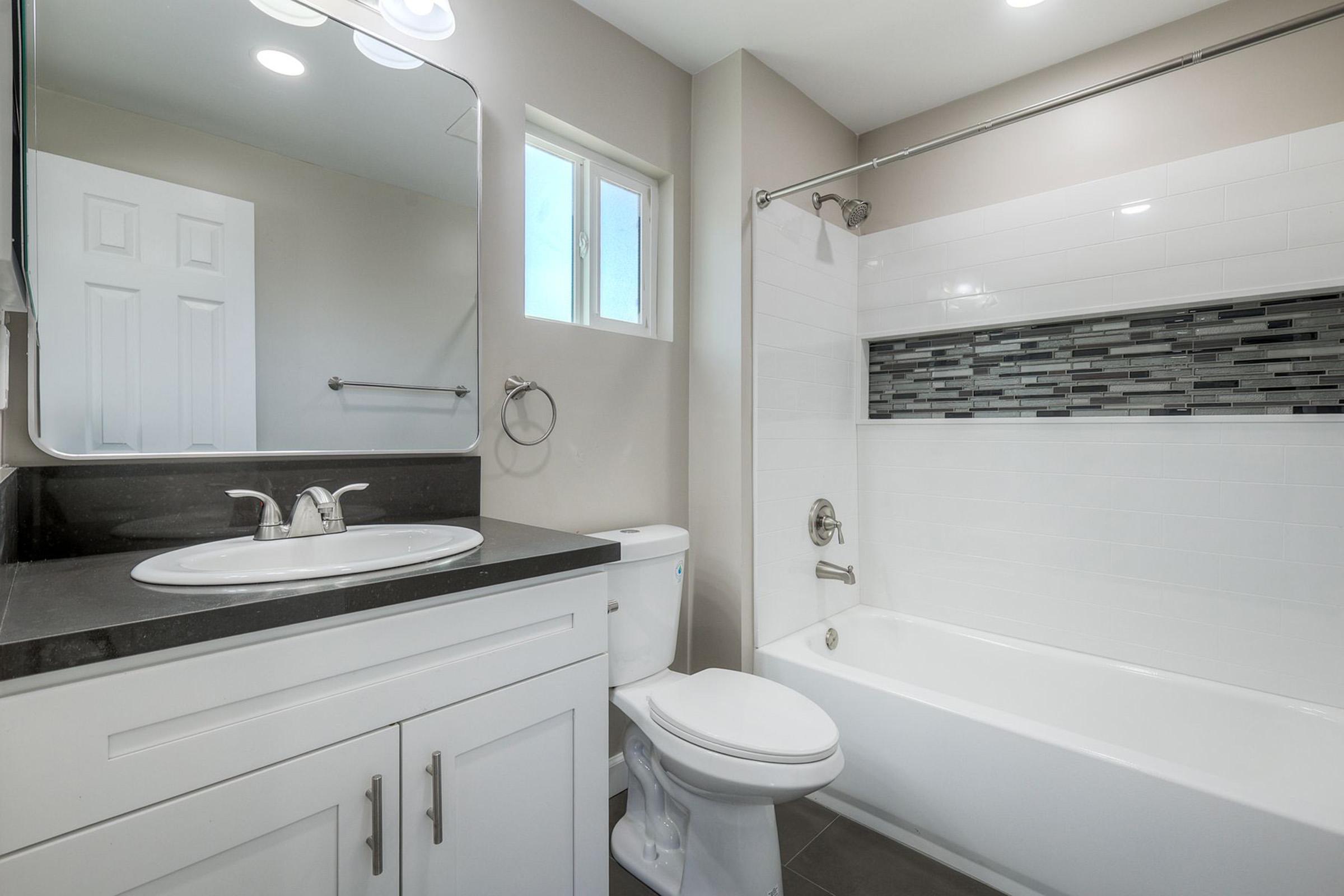A modern bathroom featuring a white bathtub with a gray and black tiled accent, a wall-mounted mirror with lighting, a white sink with a dark countertop, and a toilet. Natural light enters through a small window, complementing the neutral color scheme.