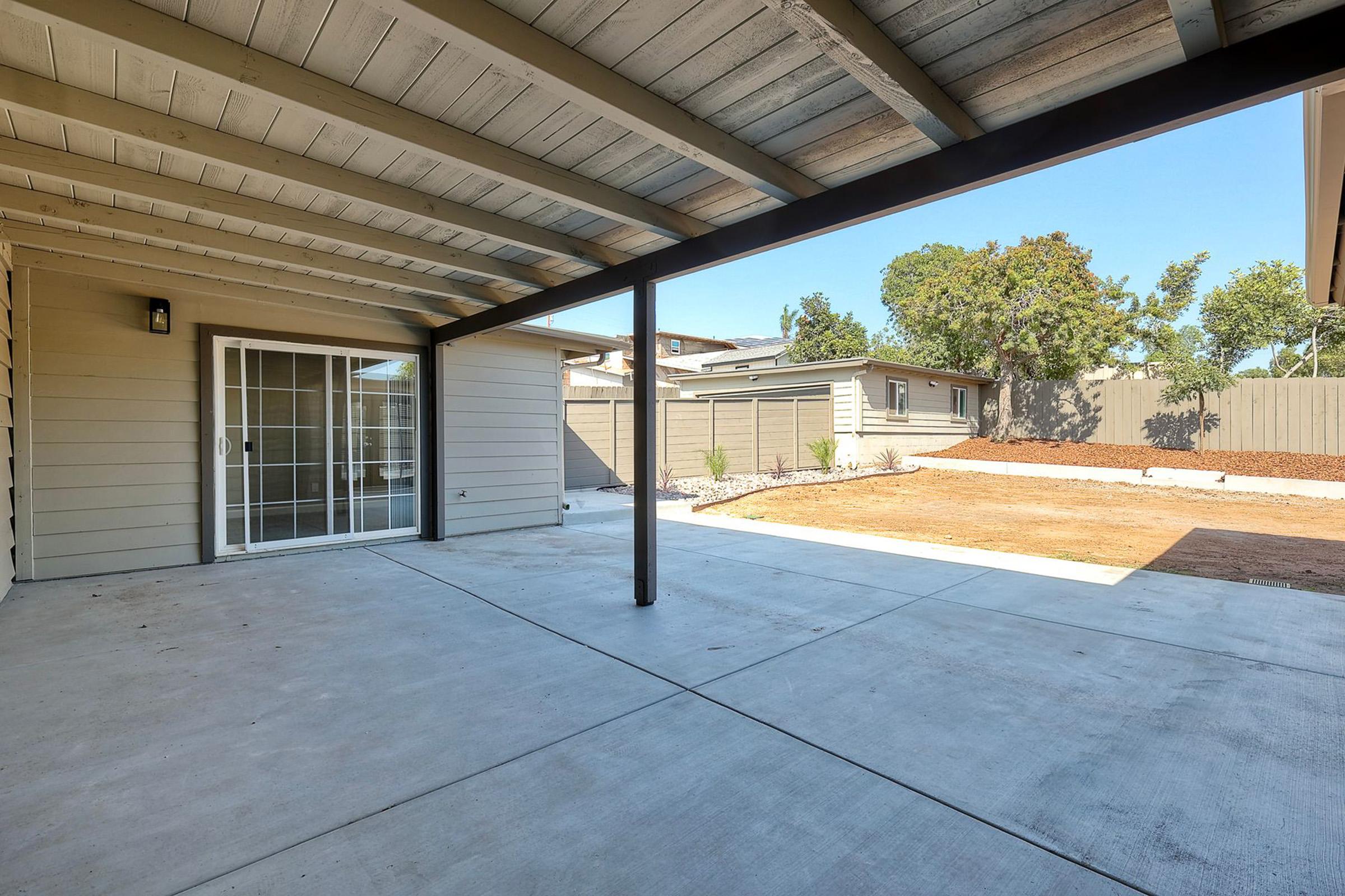 A spacious covered patio with a concrete floor, featuring a sliding glass door leading to the interior of a home. In the background, there is a fenced yard with freshly cleared land and scattered trees, under a clear blue sky. The scene conveys a bright and open outdoor living space.