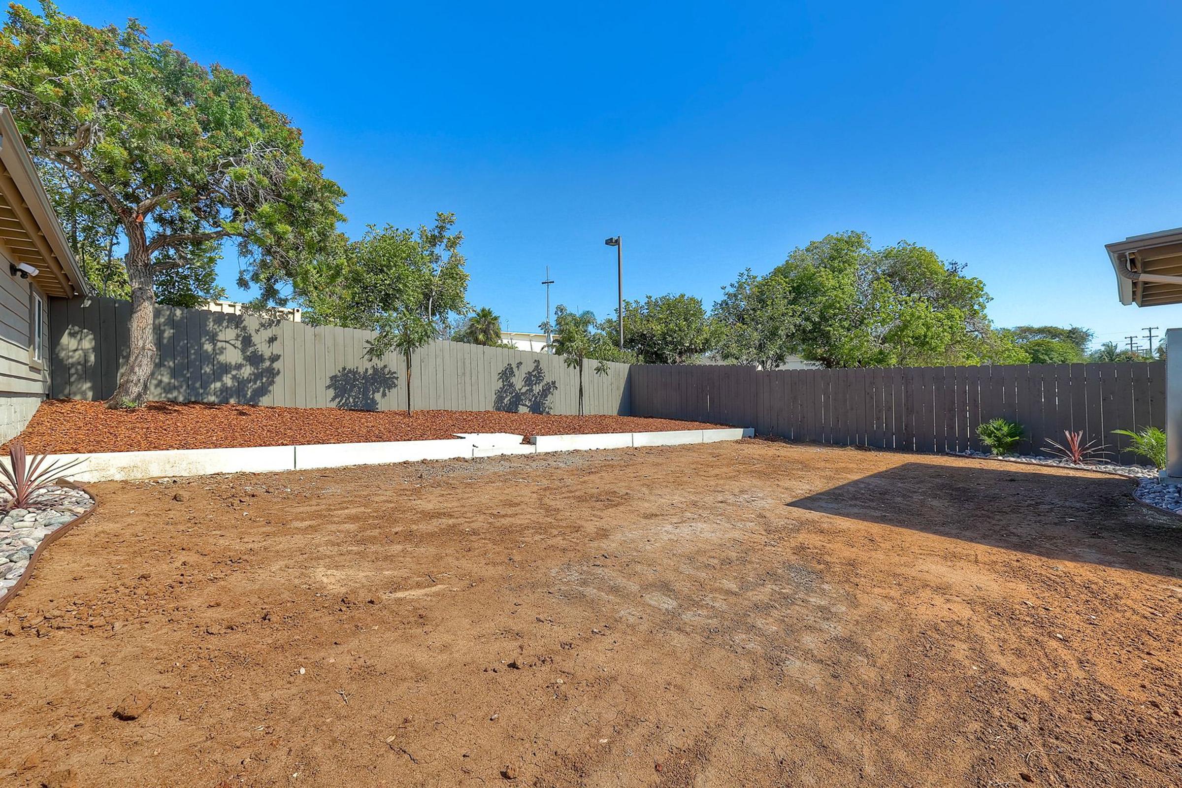 A spacious, empty backyard featuring a dirt surface and minimal vegetation, surrounded by trees and a wooden fence. The area is ready for landscaping or other outdoor projects, with clear blue skies above.