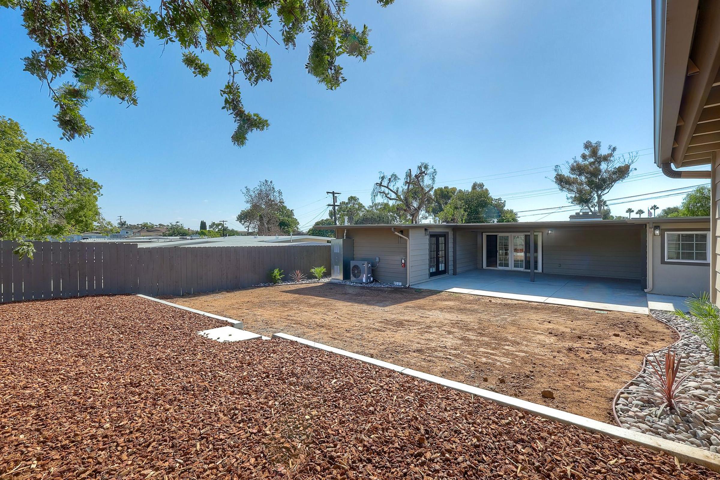 A spacious backyard with a recently landscaped area featuring mulch and a concrete patio. The space is surrounded by trees and shrubs, with a clear blue sky above. A modern home is visible in the background, with large sliding glass doors leading into the interior.