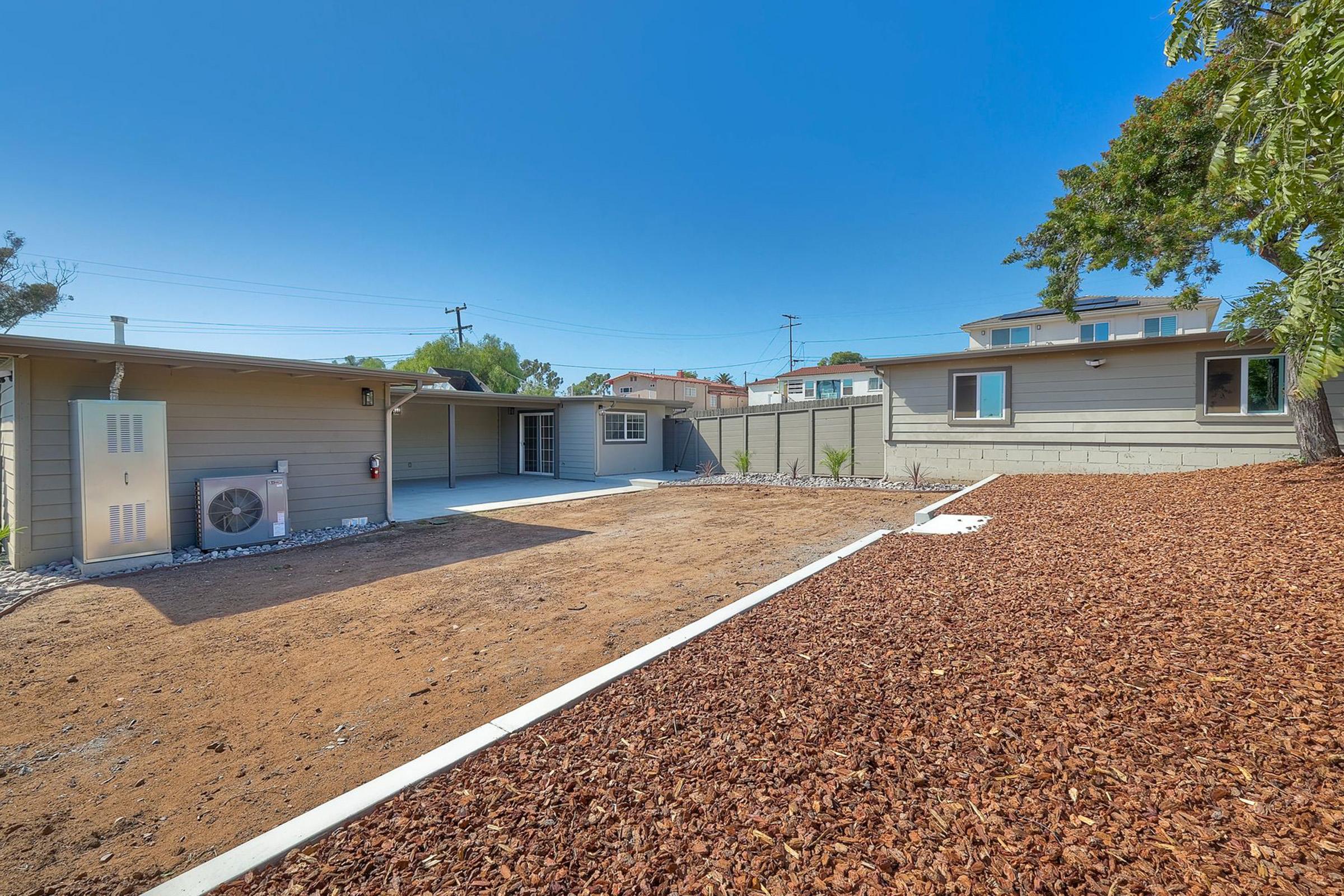 A spacious backyard with a dry, landscaped area featuring brown mulch, surrounded by a low fence. A single-story home is visible in the background, with an air conditioning unit on the side. Clear blue sky overhead, and some greenery is present around the property, indicating a sunny day.