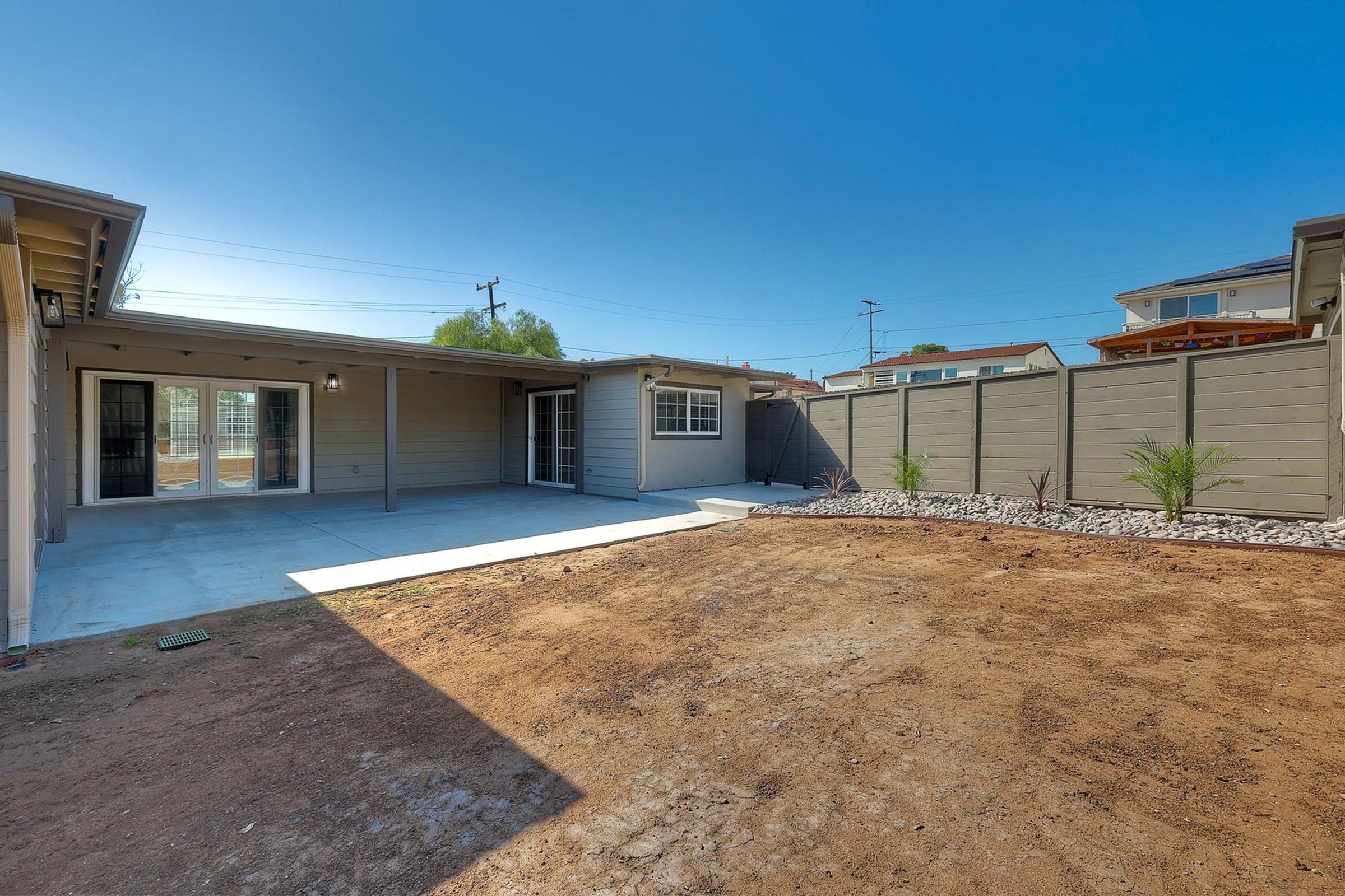 A spacious backyard area featuring a concrete patio and bare soil, surrounded by a low gray fence. The sky is clear and blue, with minimal landscaping visible. A doorway visible leads into the interior of a house, reflecting a modern design.