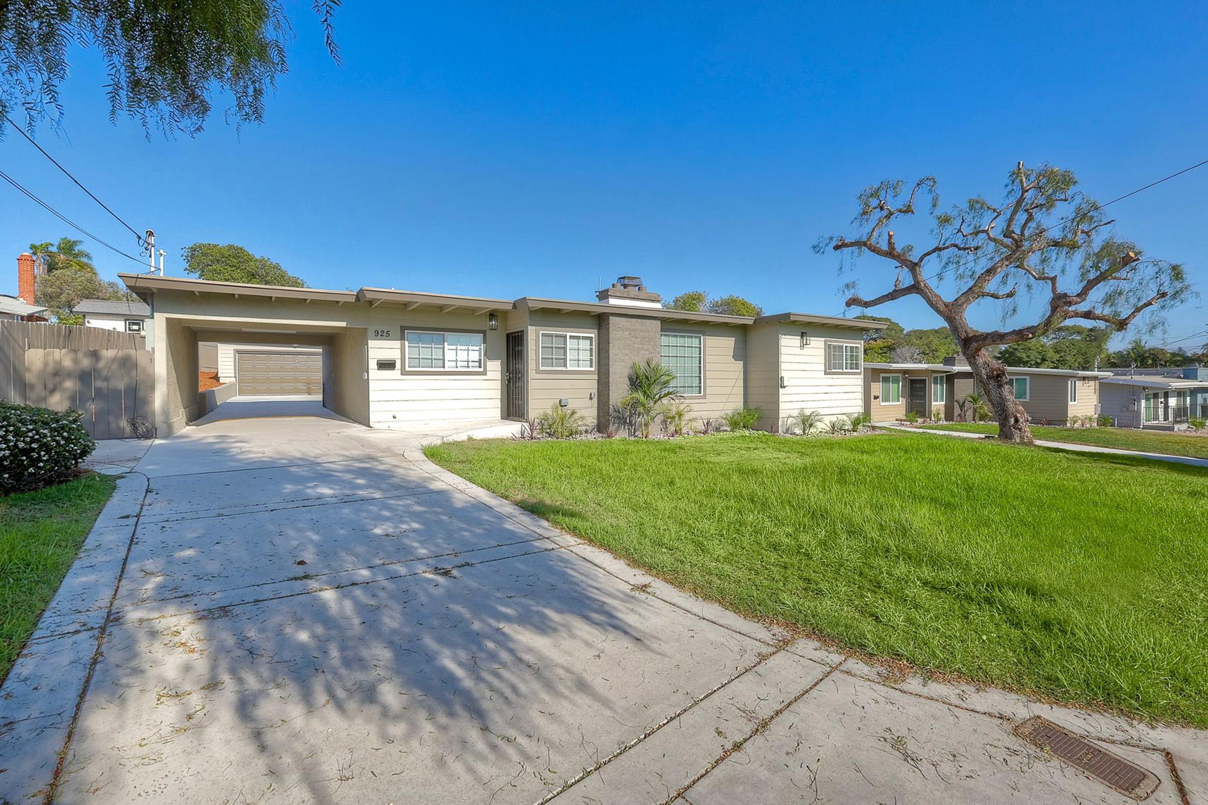 A single-story residential home with a driveway leading to a garage. The house features a light-colored facade, a chimney, and several windows. In front, there is a green lawn and a tree with sparse leaves. The sky is clear and blue, indicating a sunny day. The surrounding area includes another house in the background.