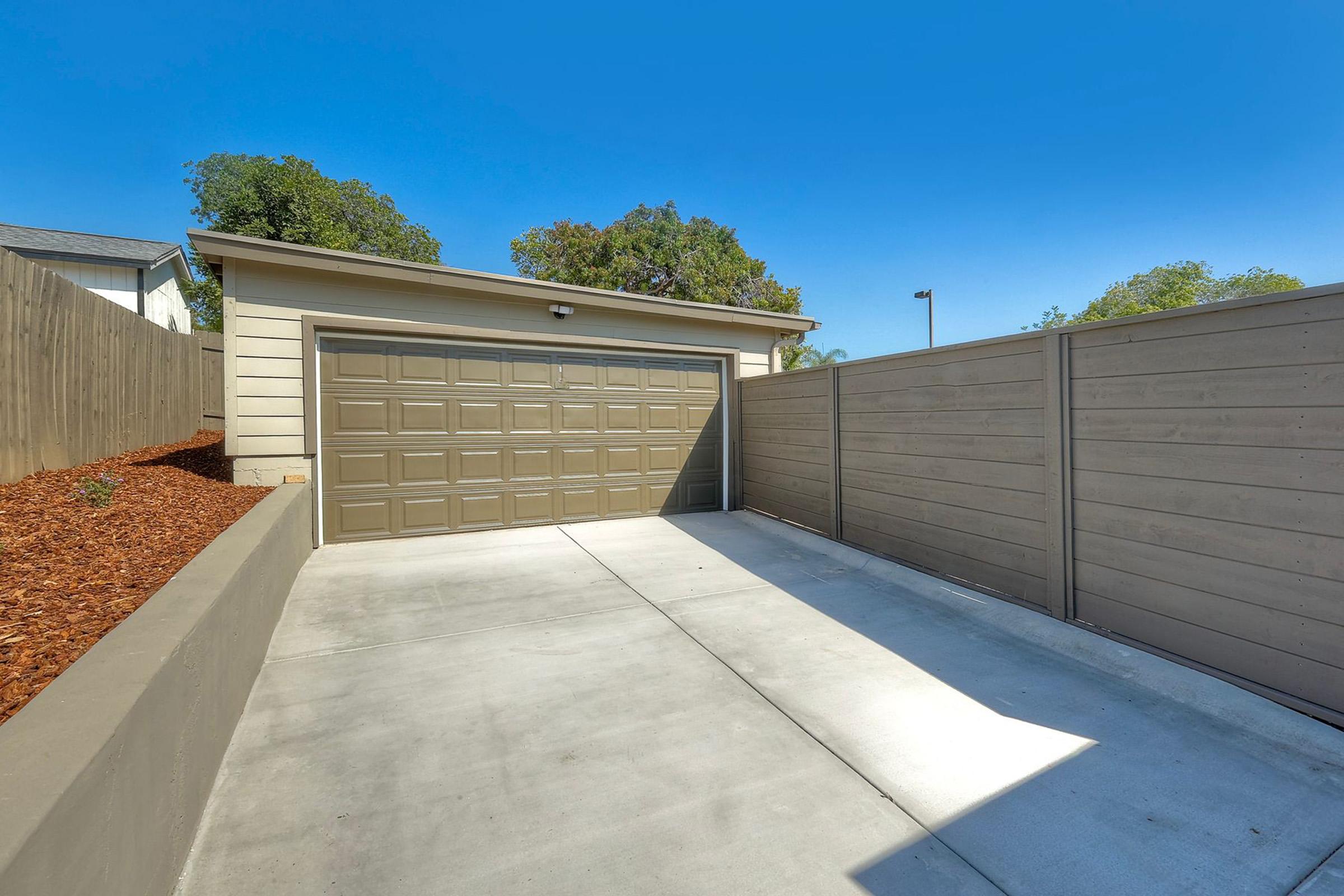 A spacious garage with a closed brown door, situated next to a gravel-covered area and flanked by a tall, modern gray fence. The clear blue sky overhead adds brightness to the scene, highlighting the clean concrete driveway leading up to the garage.