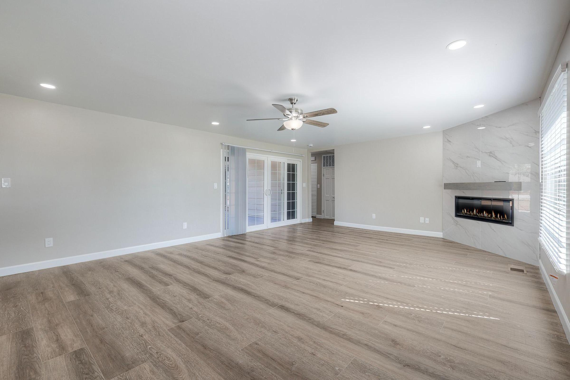 A spacious living room featuring light-colored walls, a ceiling fan, and large windows. The wooden floor complements a modern gas fireplace on one side. There are glass double doors leading to another room, creating an airy atmosphere with plenty of natural light.