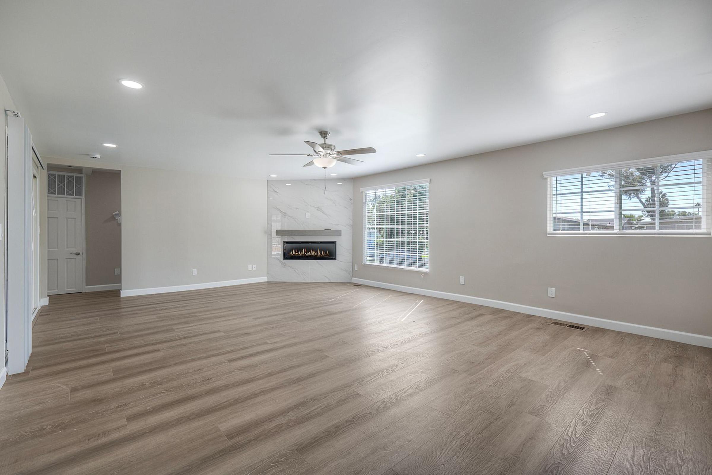 Spacious living room featuring light-colored walls, modern ceiling fan, and large windows allowing natural light. A minimalist fireplace is set against a stylish accent wall, with sleek, light hardwood flooring throughout the area.