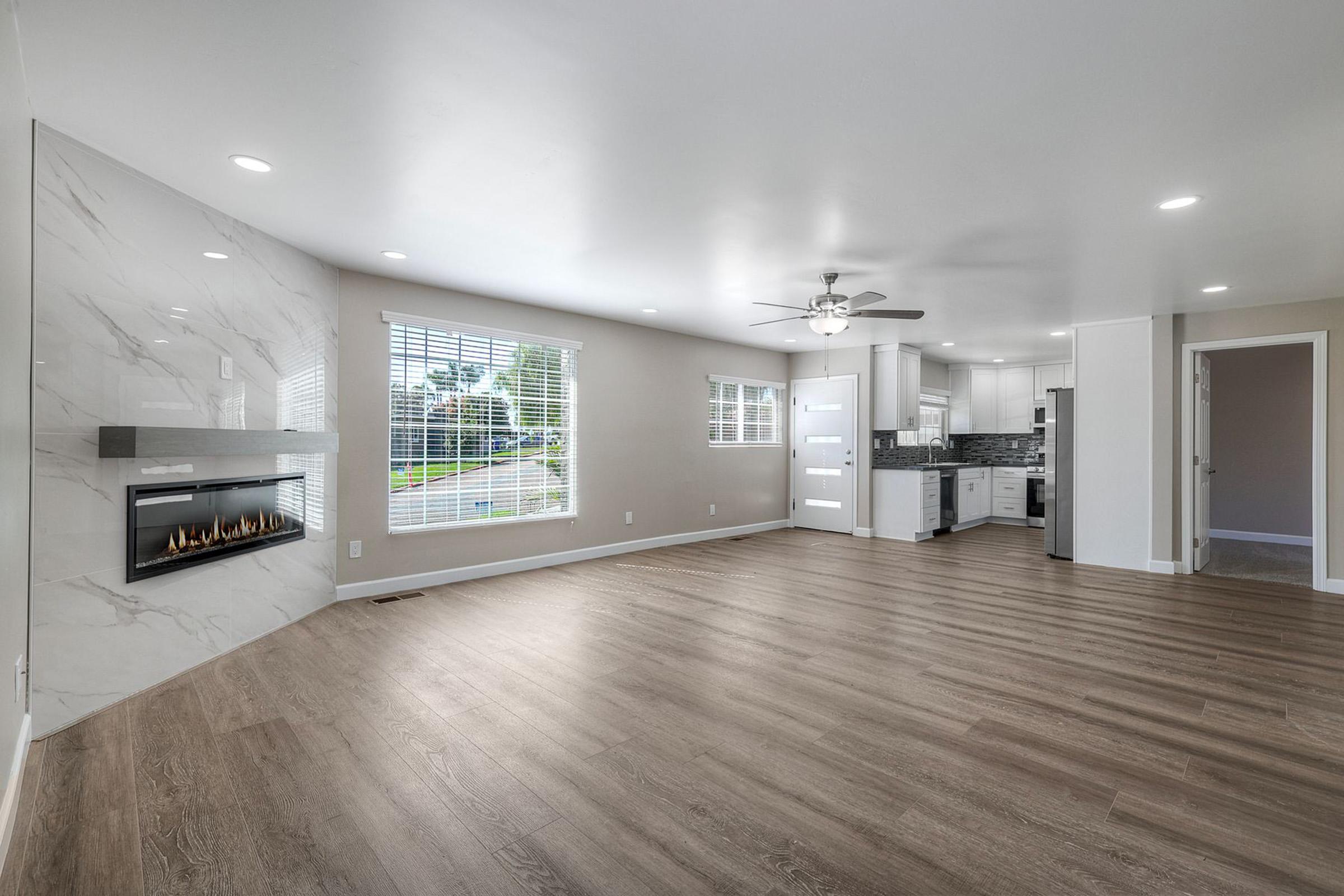 Spacious living room with modern design featuring a sleek fireplace on a marble accent wall, large windows allowing natural light, and an open layout connecting to a bright kitchen. The floor is finished with light-colored wood laminate, enhancing the airy feel of the space.