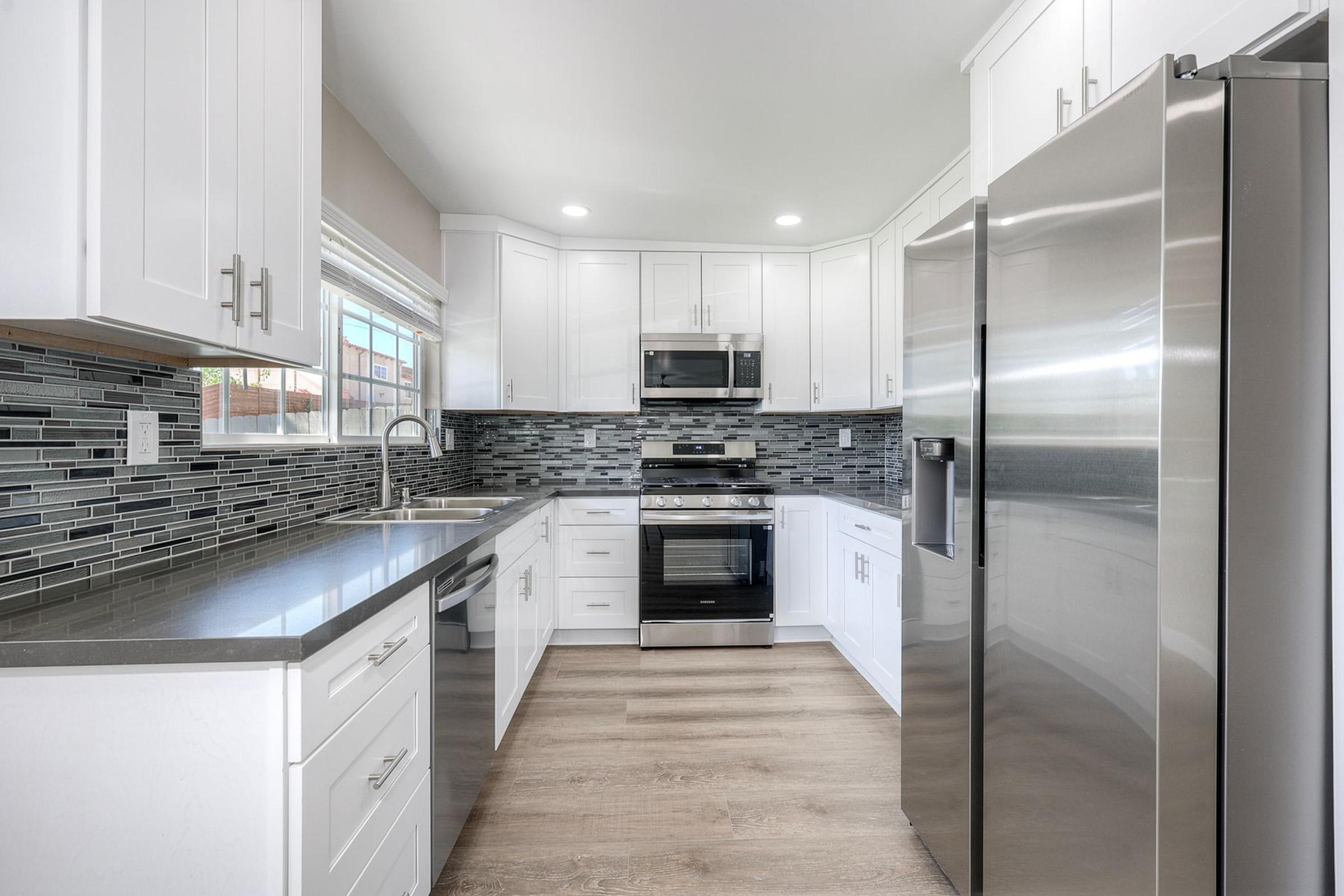 Modern kitchen featuring white cabinetry, stainless steel appliances, a stylish gray countertop, and a backsplash of mixed gray and black tiles. Abundant natural light comes through a window, illuminating the space with a clean and contemporary design.