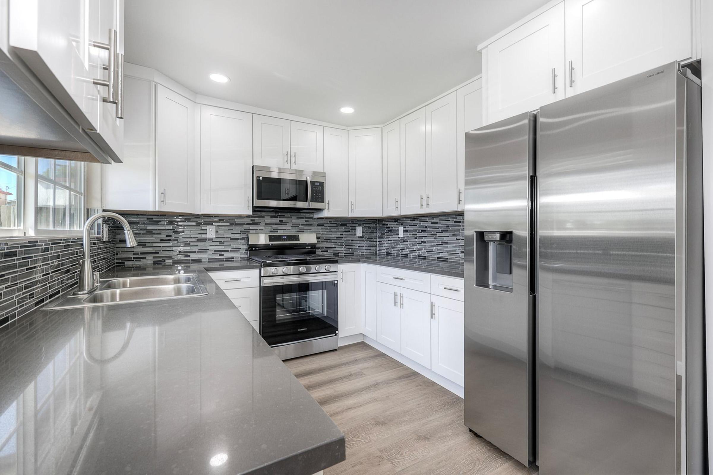 Modern kitchen featuring white cabinets, stainless steel appliances including a refrigerator and oven, a microwave mounted above the stove, and a gray and black tiled backsplash. The countertops are dark and there are dual sinks with a sleek faucet. Bright lighting enhances the spacious, clean design.