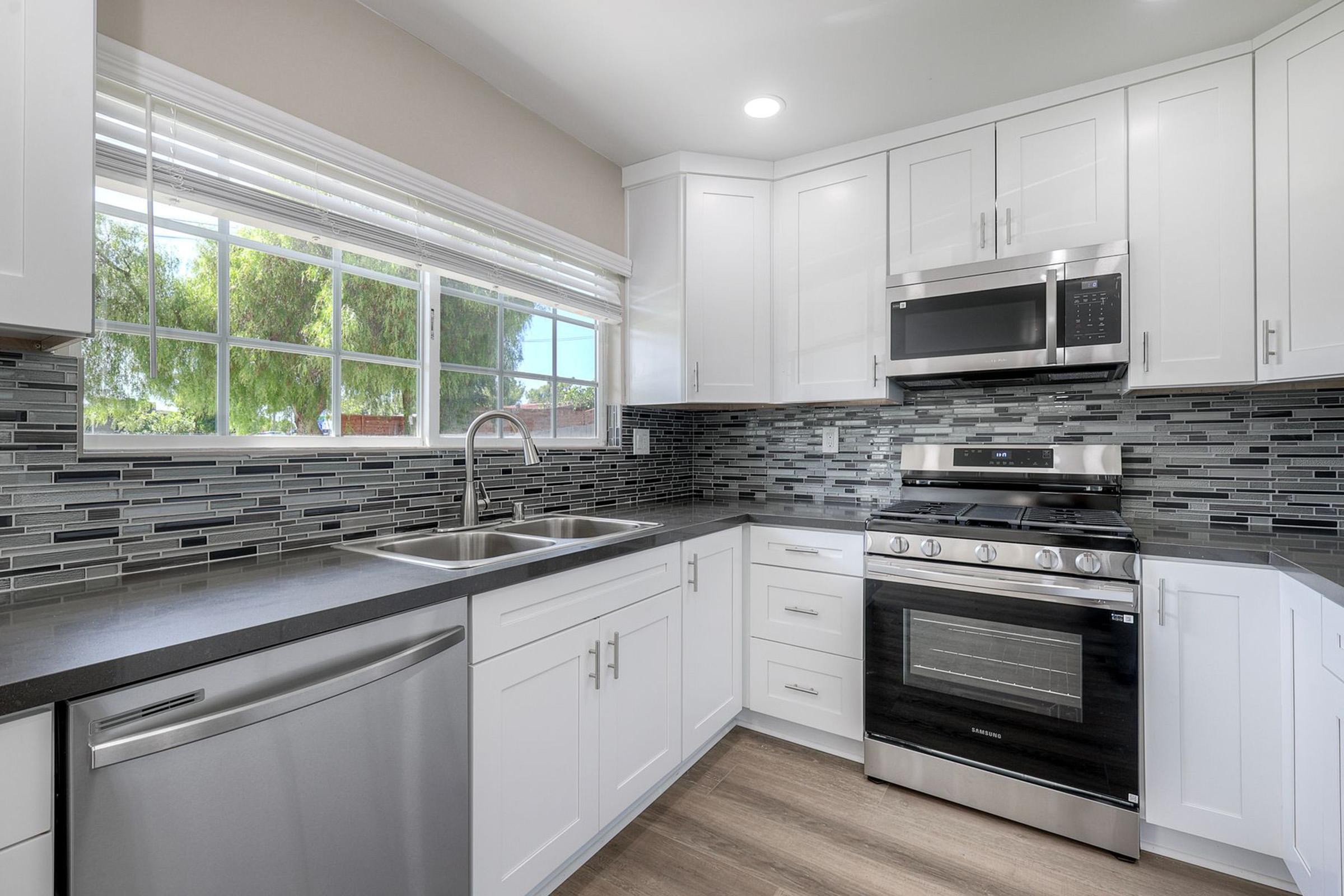 Modern kitchen featuring white cabinets, stainless steel appliances, and a gray countertop. A double sink is under a window with a view of greenery. Backsplash made of black and gray tiles enhances the contemporary design. Natural light brightens the space, creating a clean and inviting atmosphere.