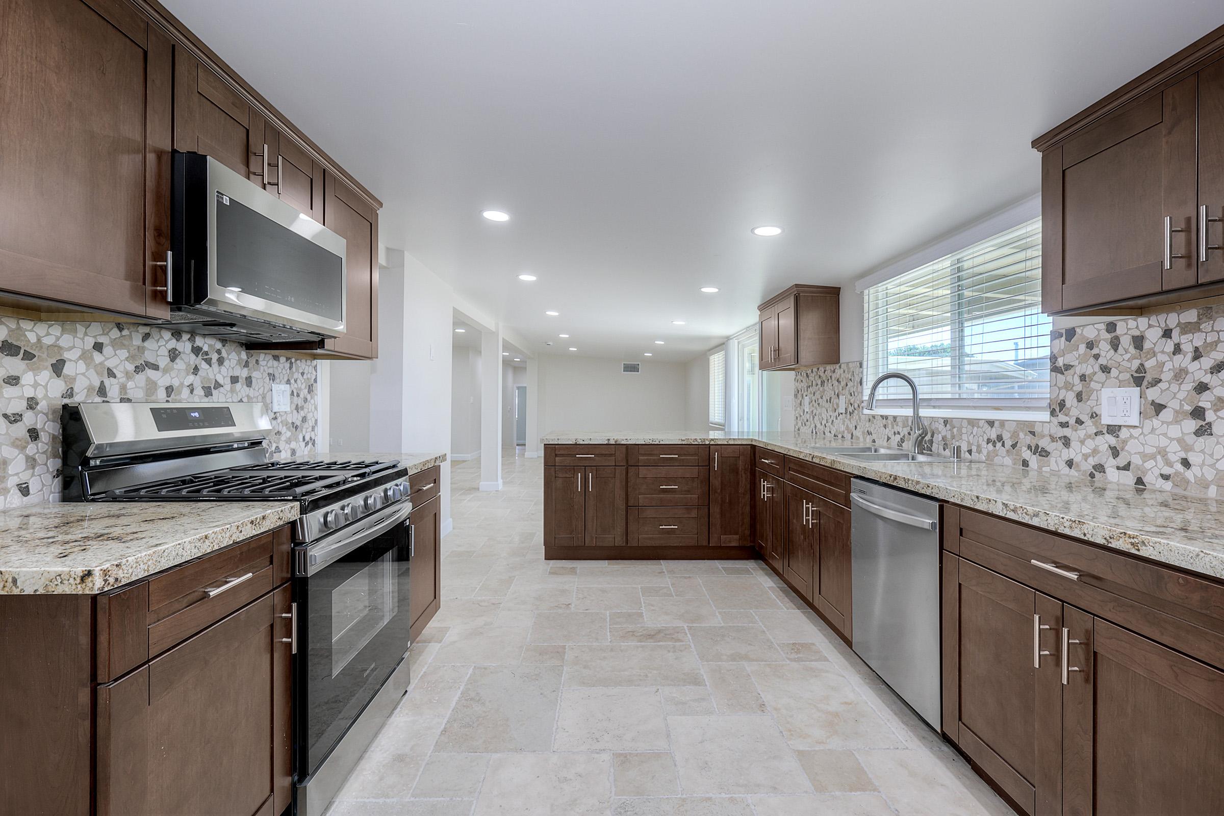 Modern kitchen featuring dark wood cabinetry, stainless steel appliances, and a granite countertop. The backsplash is made of mosaic tiles, and the space has ample natural light from windows, enhancing its bright and inviting atmosphere. The flooring consists of light-colored tiles that complement the overall design.