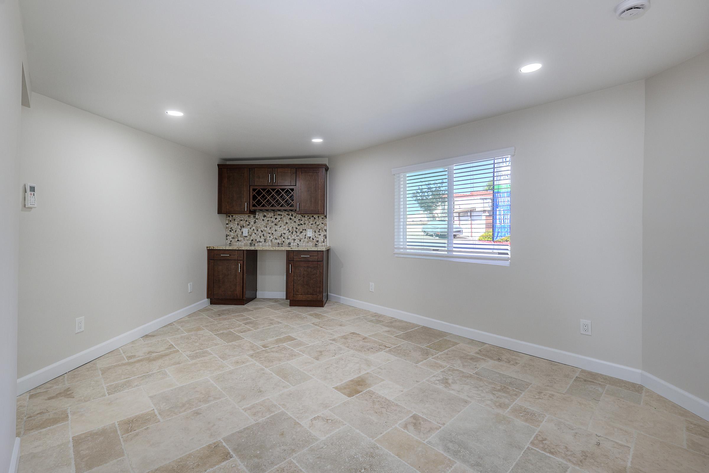 A spacious, well-lit room featuring beige tiled flooring and white walls. On one side, there is a built-in wooden cabinet with a decorative backsplash, accompanied by a window allowing natural light to enter. The space is clear of furniture, emphasizing its openness.