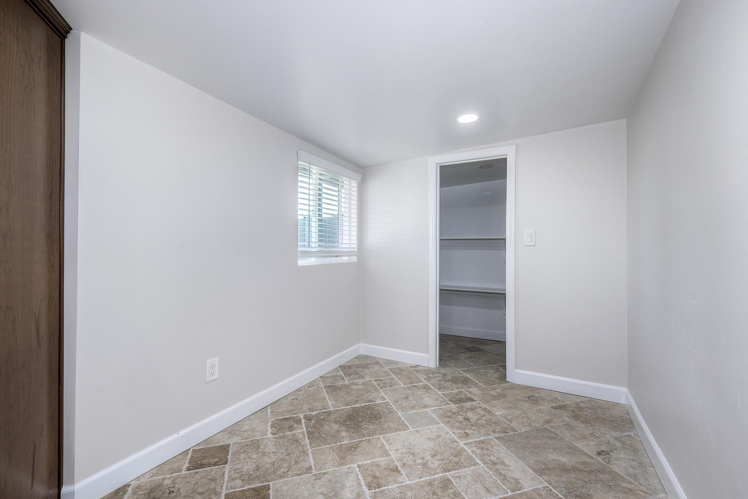 A clean, empty room with light-colored walls and tiled flooring. A small window with blinds allows natural light in, and there is a door leading to a closet or storage space. The room appears well-lit and spacious, suitable for various purposes.