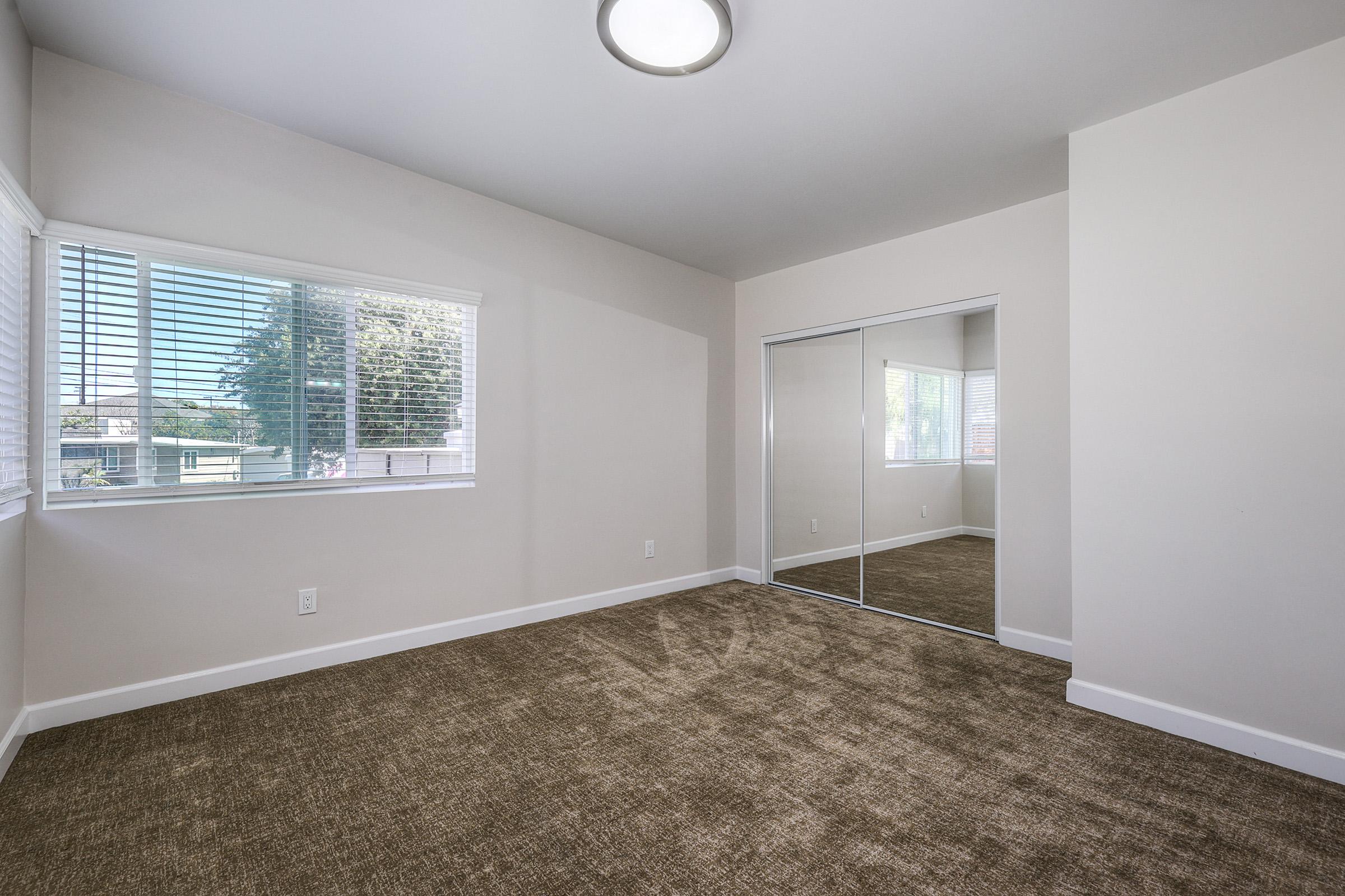 A light-filled, empty bedroom featuring a large window with blinds, beige walls, and a carpeted floor. A mirror closet reflects the room, adding depth to the space. The overall atmosphere is clean and modern, ideal for personal decoration.