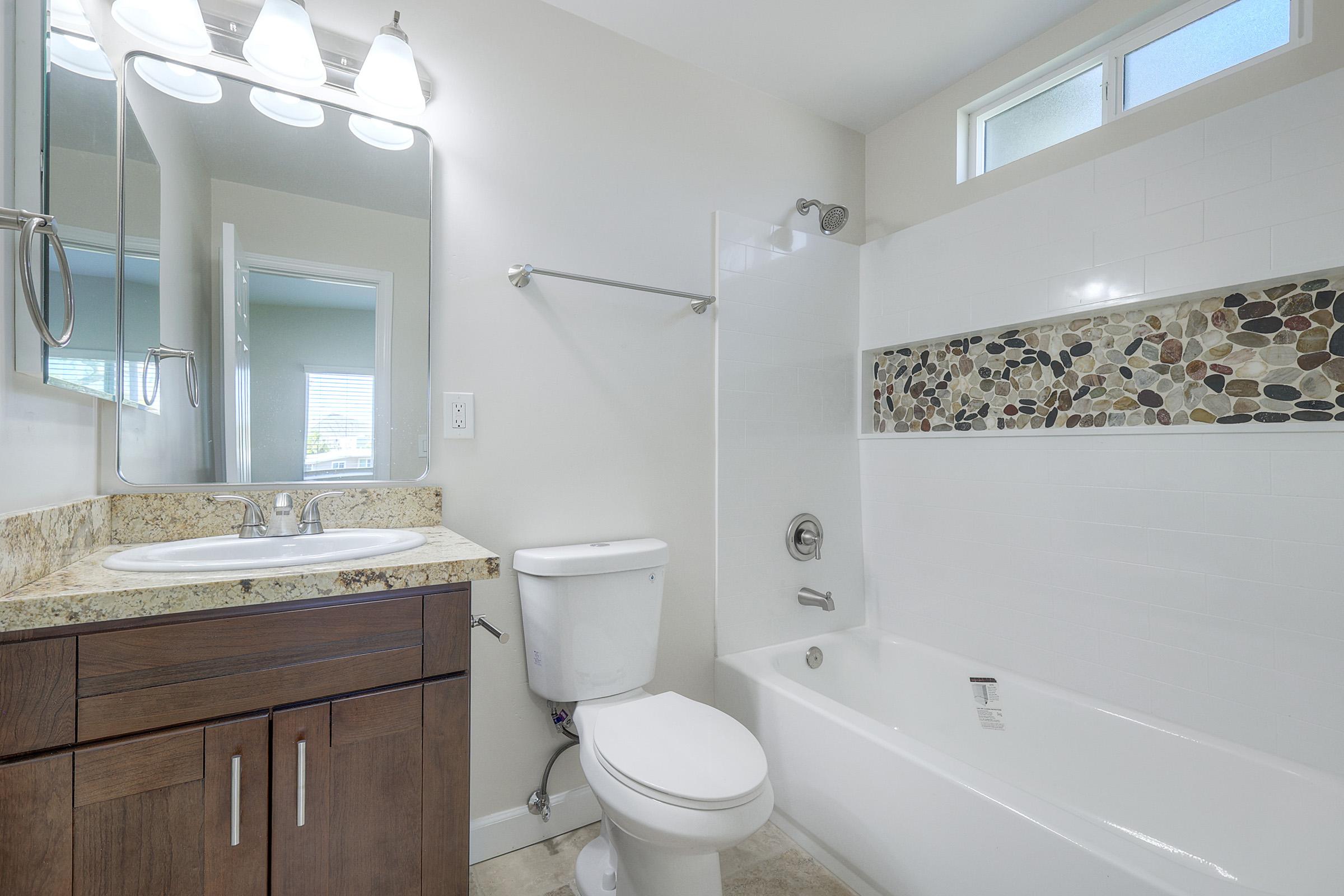 A modern bathroom featuring a bathtub with a decorative stone accent, a toilet, a stone countertop sink with a mirror above, and a bright, airy atmosphere enhanced by natural light from a window. The walls are painted in a neutral tone, creating a clean and spacious look.