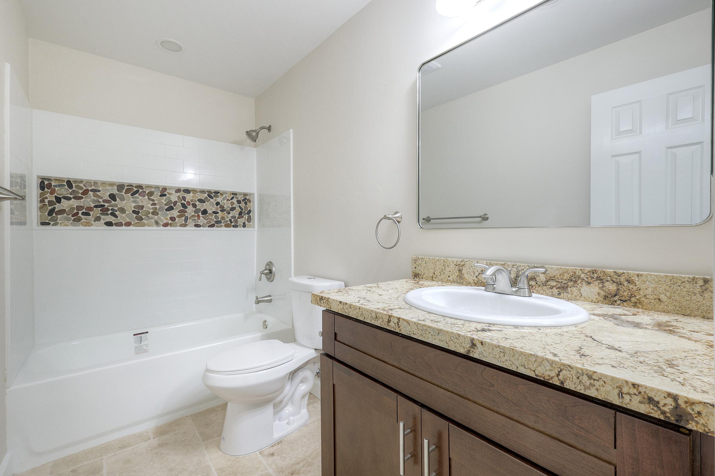 A modern bathroom featuring a white bathtub with a decorative stone accent wall, a wooden vanity with a granite countertop, a round sink, a mirror above the sink, and a tiled floor. The walls are painted in neutral tones, and there's a towel rack next to the toilet.