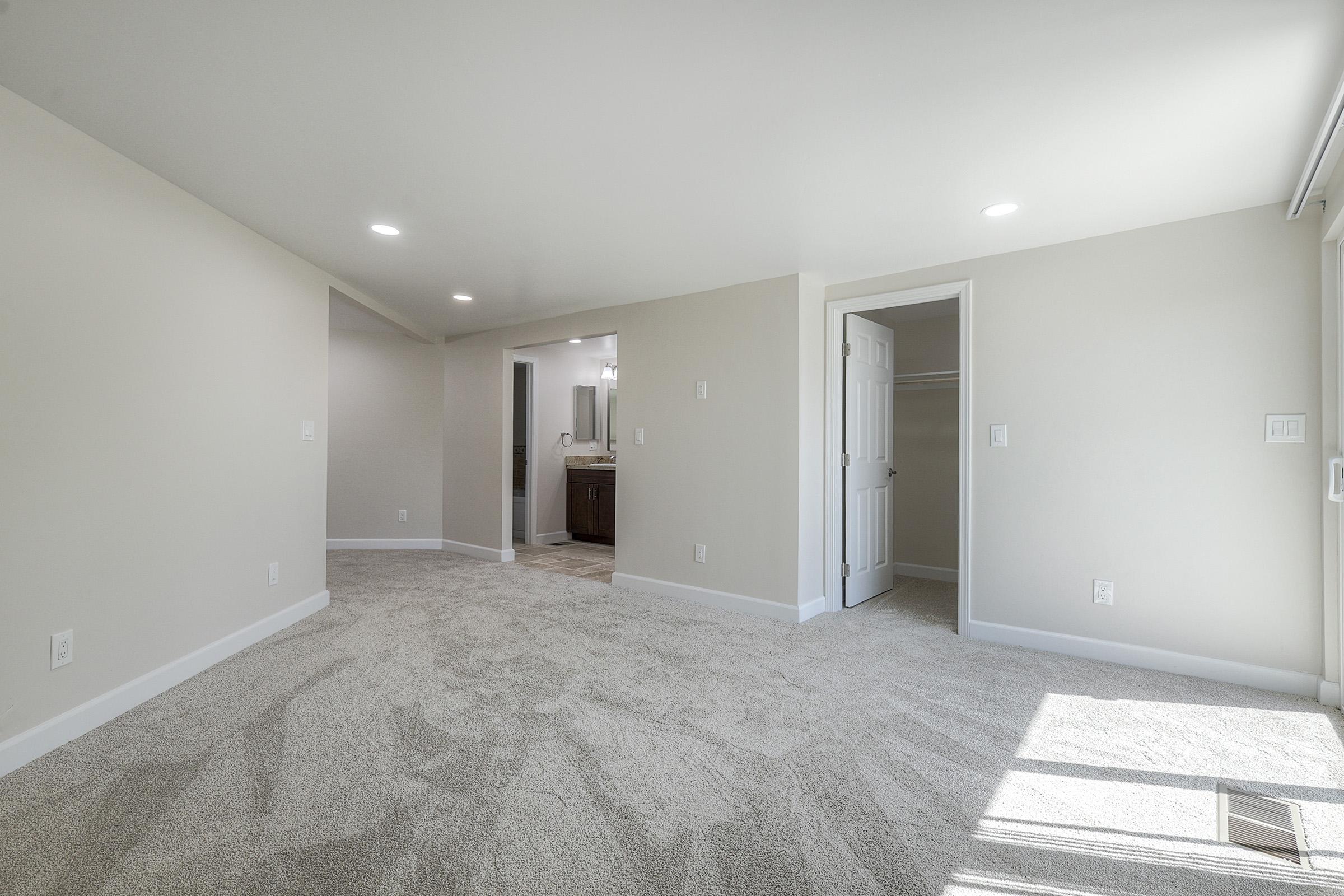 A spacious and well-lit interior room with neutral-colored walls, plush carpet, and recessed lighting. An open doorway leads to a bathroom area, while a closet is visible on the right. Large windows allow natural light to filter in, creating a bright and inviting atmosphere.