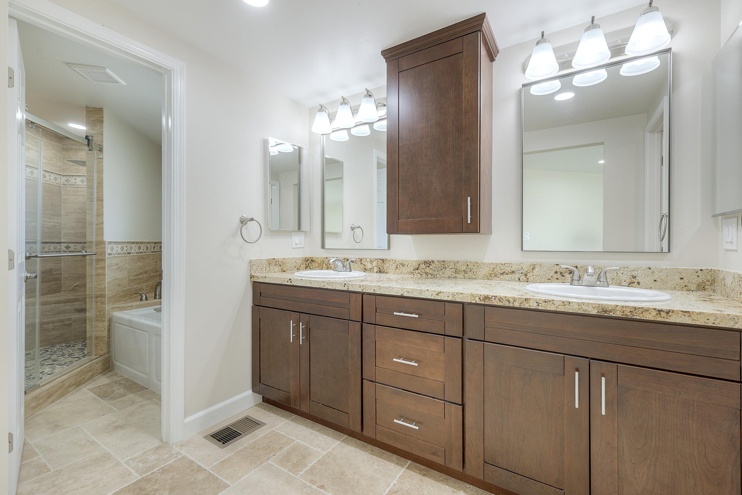 A modern bathroom featuring a double vanity with wooden cabinetry and two sinks, illuminated by wall-mounted light fixtures. The walls are painted in a light color, and there are two mirrors above the sinks. A shower area with tile designs is visible in the background, along with a separate tub.