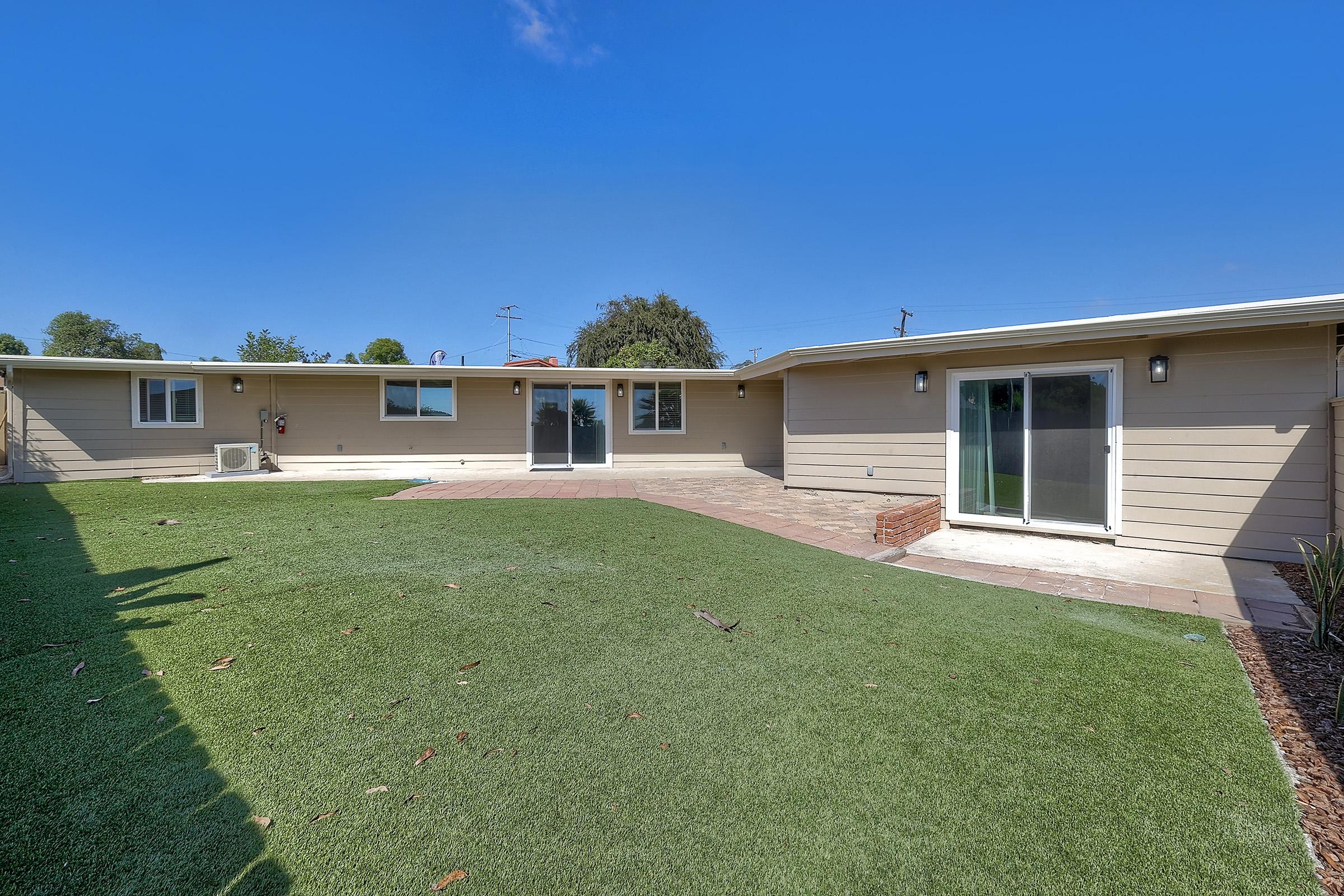 A backyard view of a single-story home featuring a well-manicured lawn with artificial grass. There is a patio area with stone paving and sliding glass doors leading into the home. The sky is clear and blue, making the space appear bright and inviting. Trees and power lines are visible in the background.