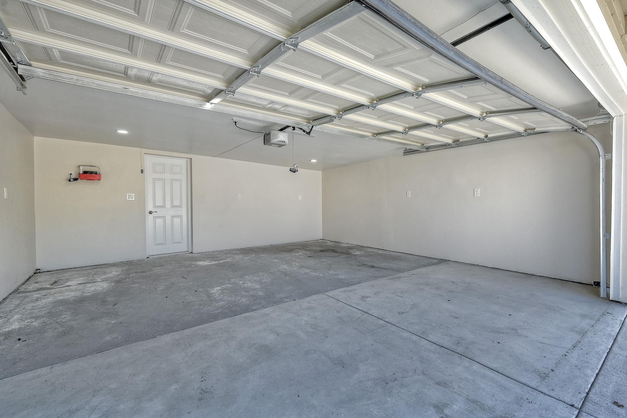 Empty garage interior featuring a concrete floor, plain white walls, and a ceiling with a multi-panel door. There is a closed door on one wall and a small light fixture on the opposite wall, creating a spacious and clean environment suitable for parking or storage.