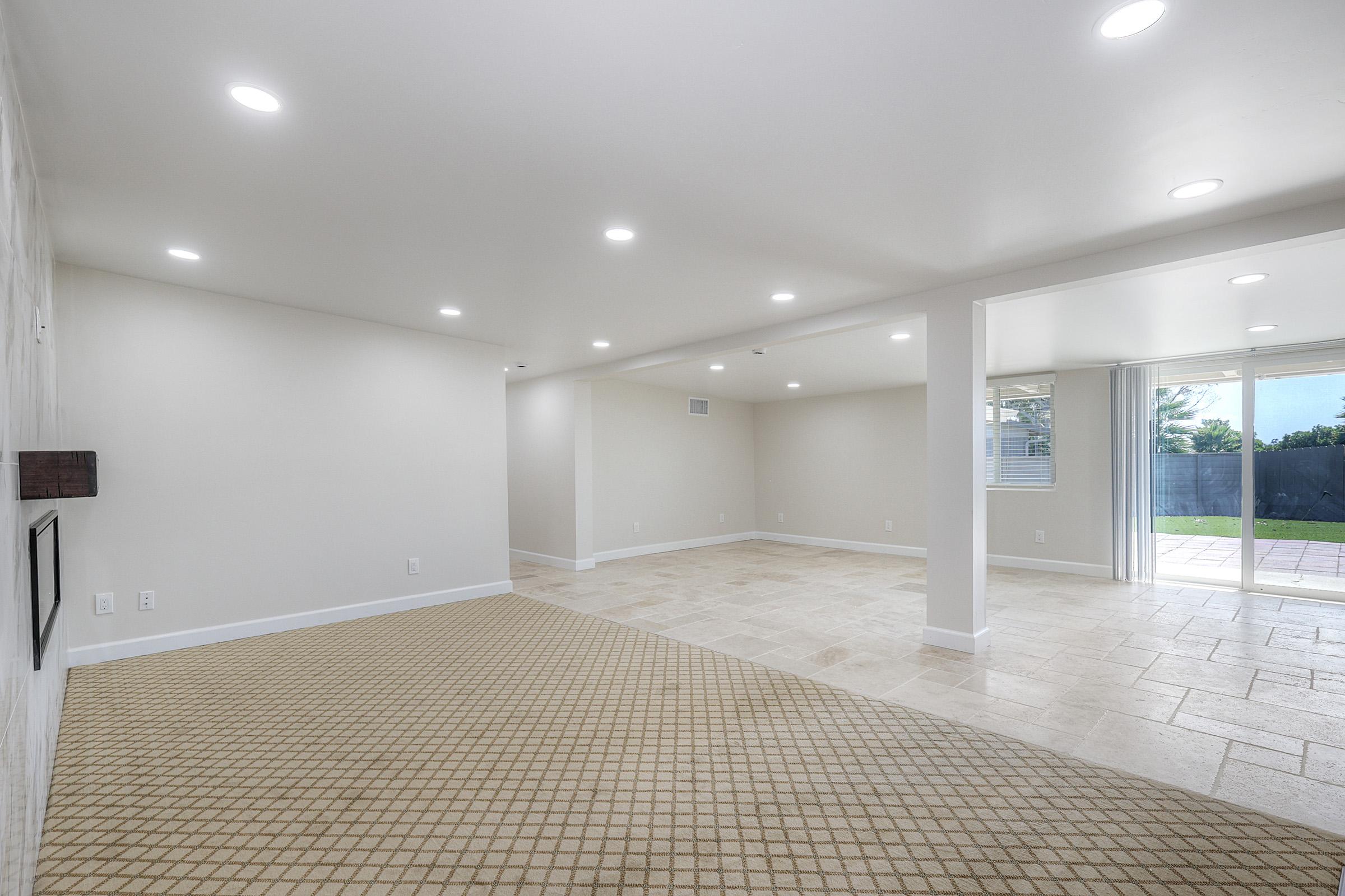 A spacious, well-lit room featuring light-colored walls, recessed lighting, and large tile flooring. One side has a textured pattern on the floor, while large glass doors open to an outdoor area. The room is empty, offering a versatile space for various uses.