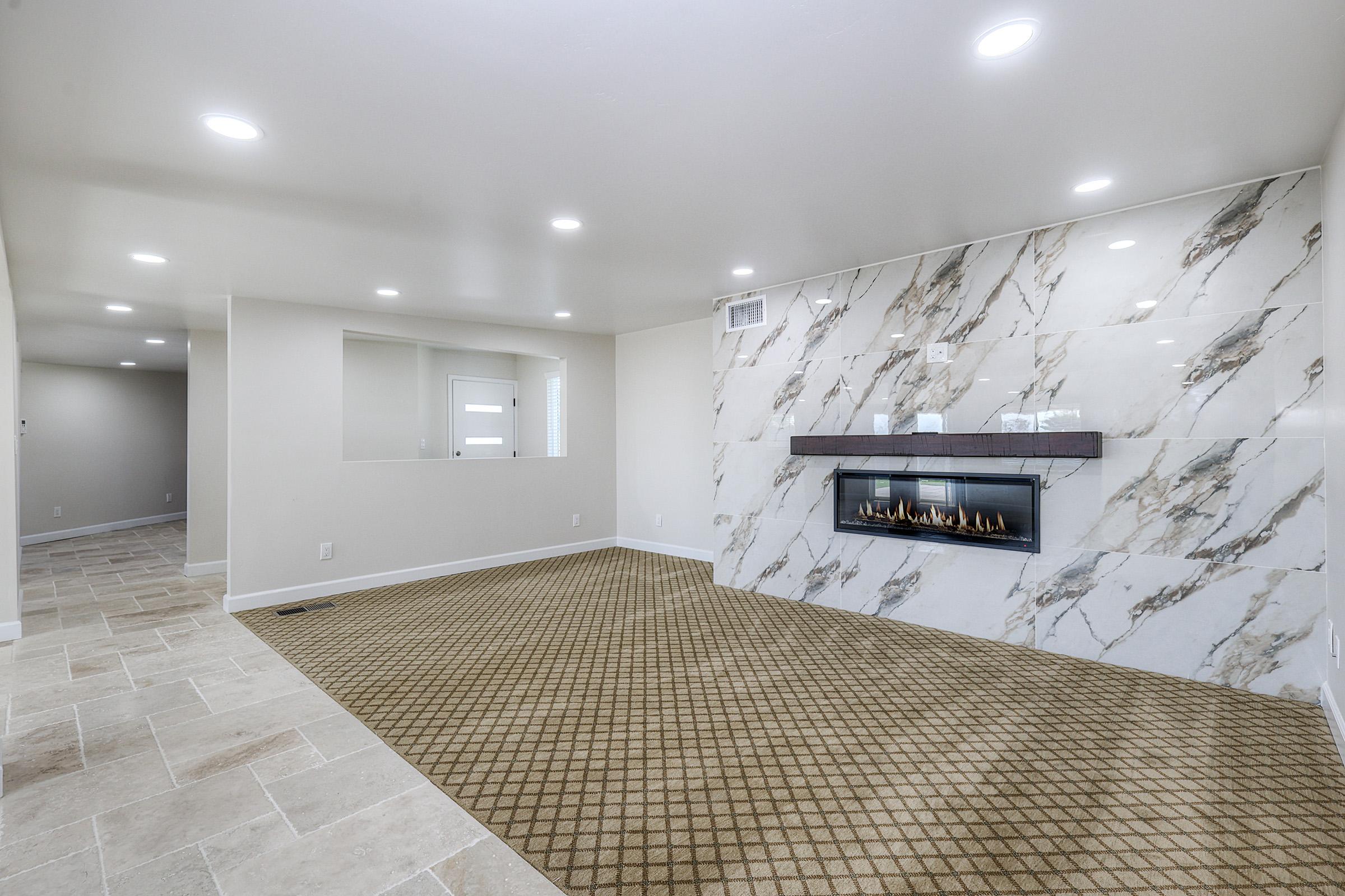 Modern living room featuring a marble accent wall with a sleek fireplace, beige walls, and a textured tan carpet. Soft ceiling lights provide illumination, and a small window adds natural light. The space is designed for comfort and contemporary style.