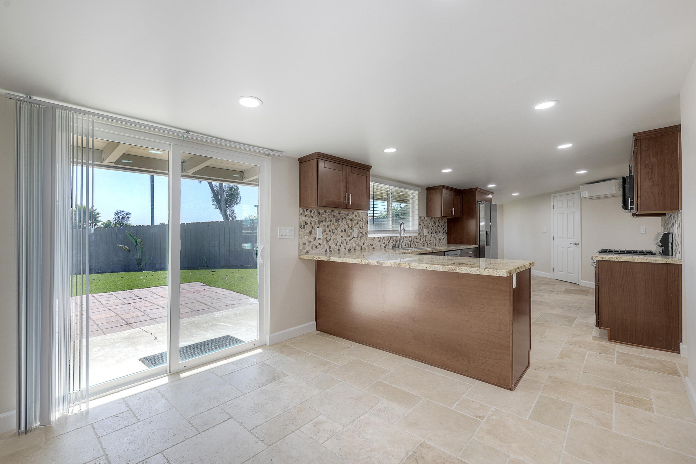 A modern kitchen and dining area featuring light-colored tile flooring, wooden cabinetry, and a spacious island with a granite countertop. Natural light floods the space through large windows, and glass sliding doors lead to a grassy outdoor area.