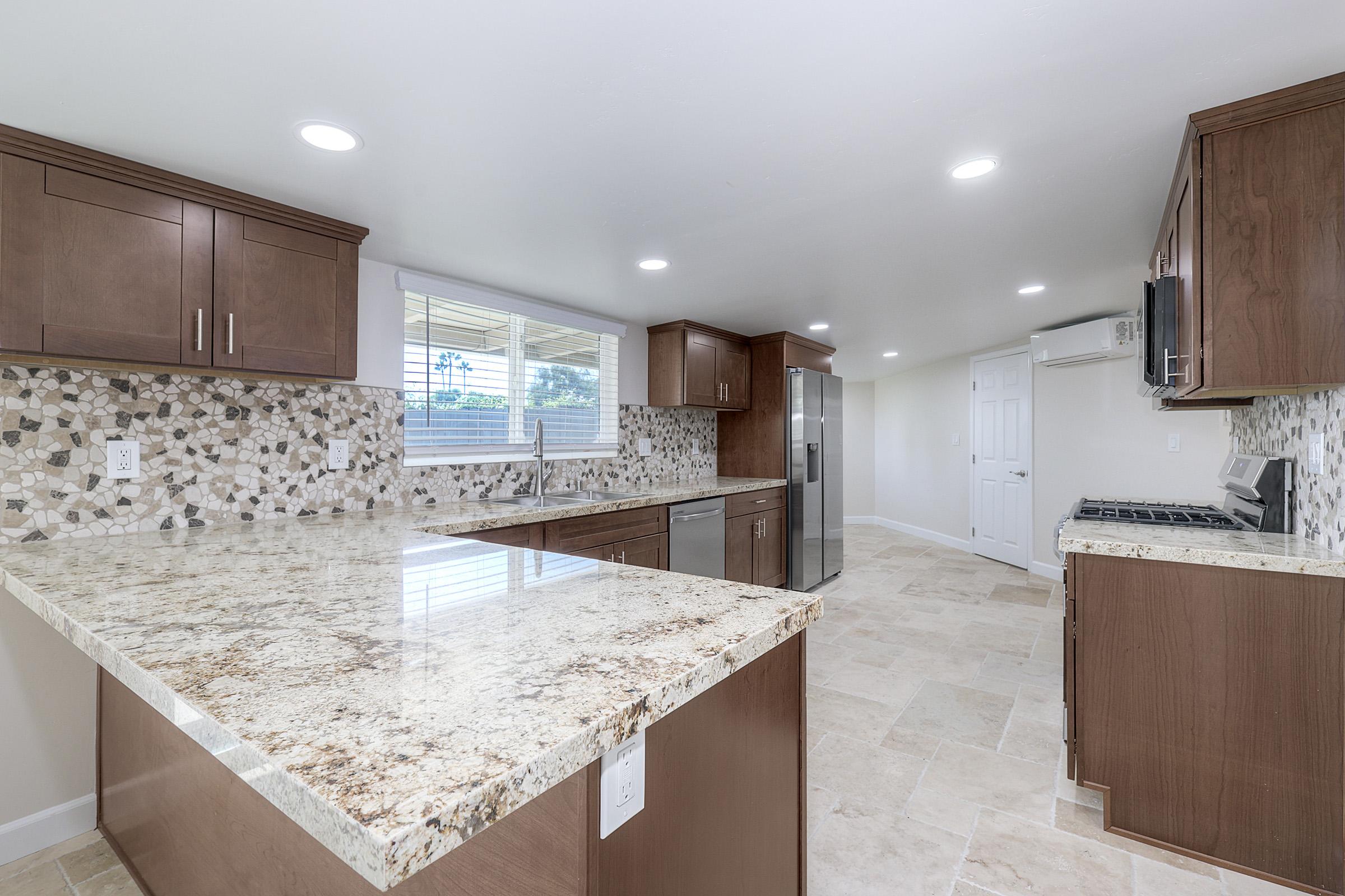 Modern kitchen featuring wooden cabinets, a large granite countertop, and a mosaic tile backsplash. Equipped with stainless steel appliances and a window allowing natural light. The floor is tiled, creating a clean and bright atmosphere in a spacious layout.
