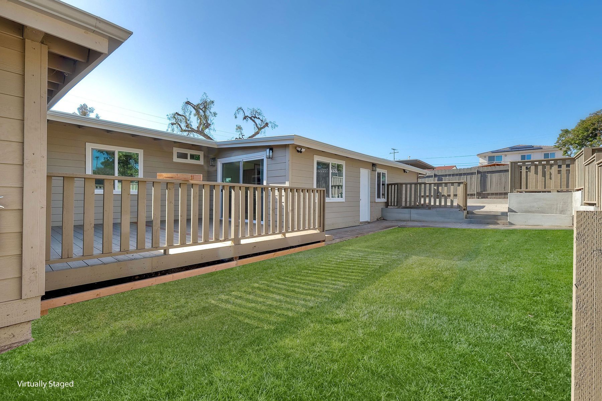 View of a backyard with a green lawn, featuring a wooden deck and a patio area. The setting includes two adjacent houses with large windows, showcasing a sunny day with clear blue skies. The area is separated by a fence, enhancing privacy. There are trees in the background, adding to the pleasant outdoor vibe.