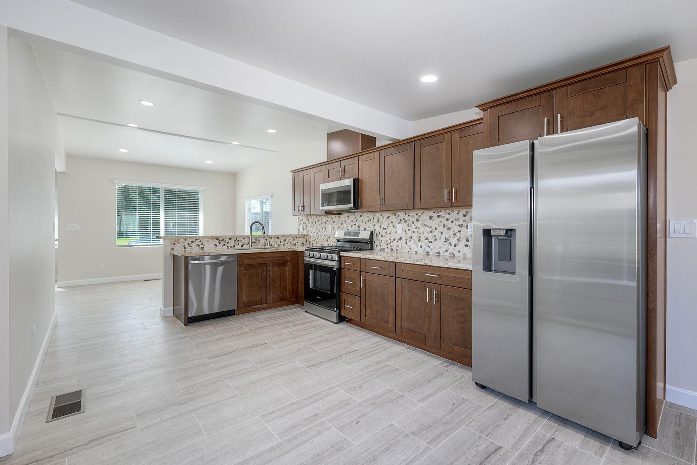 A modern kitchen featuring brown wooden cabinets, a stainless steel refrigerator, a microwave, and a gas stove. The backsplash is adorned with decorative tiles, and the floor has light-colored tiles. Natural light streams in from a nearby window, creating a bright and open atmosphere.