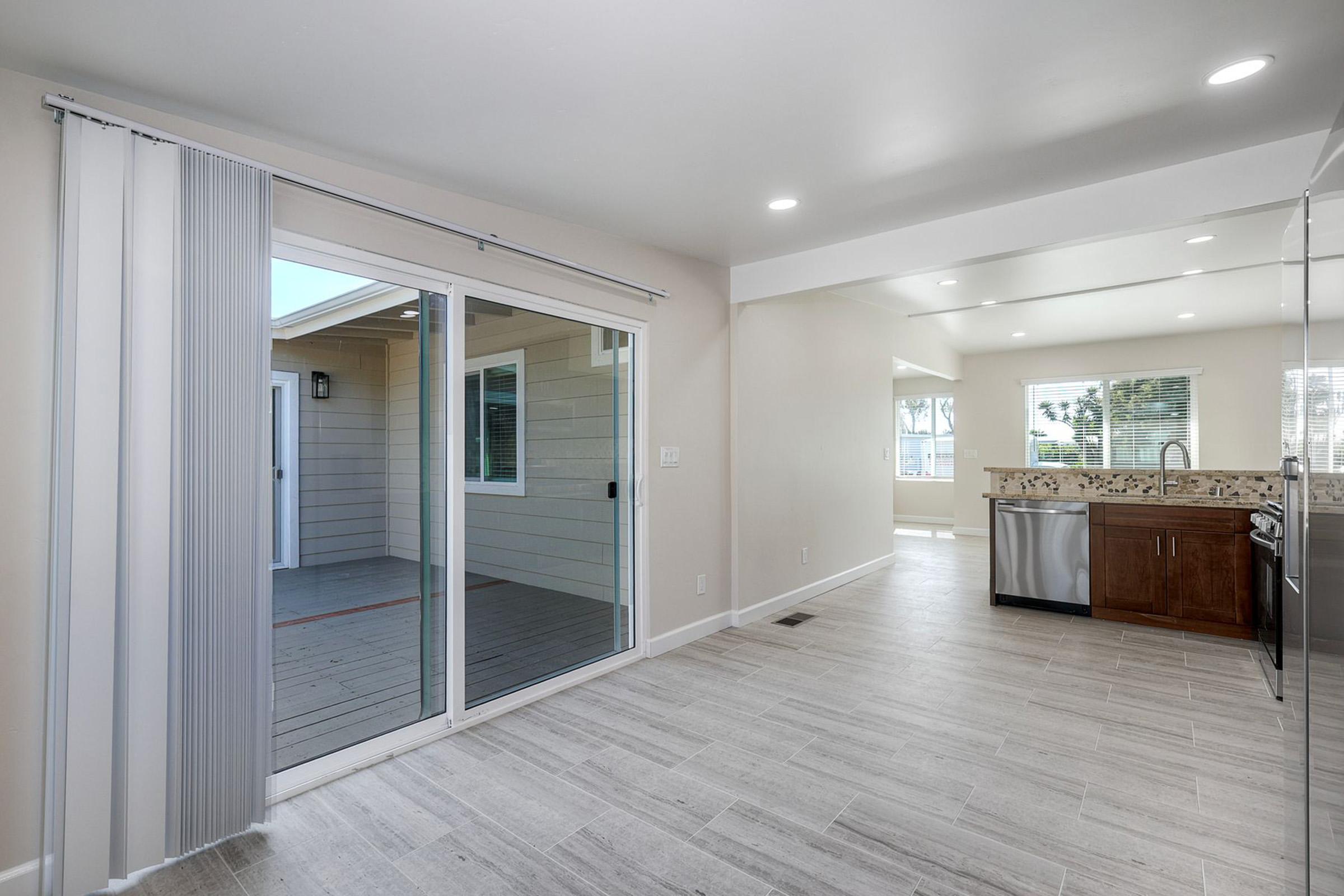 An open interior space featuring a light-colored kitchen with stainless steel appliances, a tile floor, and a large sliding glass door leading to an outdoor area. Natural light fills the room, showcasing a modern design with smooth walls and a neutral color palette.