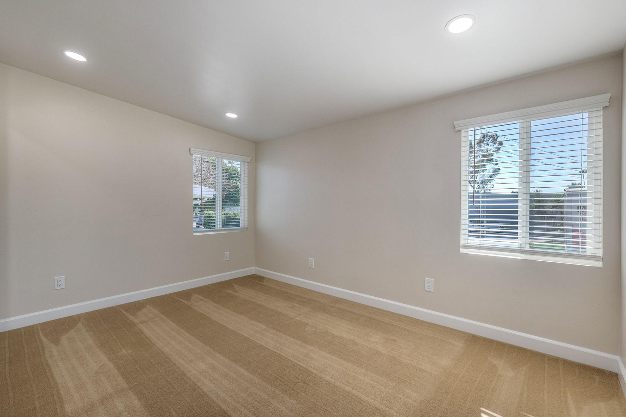 Empty room with neutral-colored walls and light-colored carpet. Two windows with blinds allow natural light, illuminating the space. The room is unfurnished, providing a feeling of openness and potential for personalization.