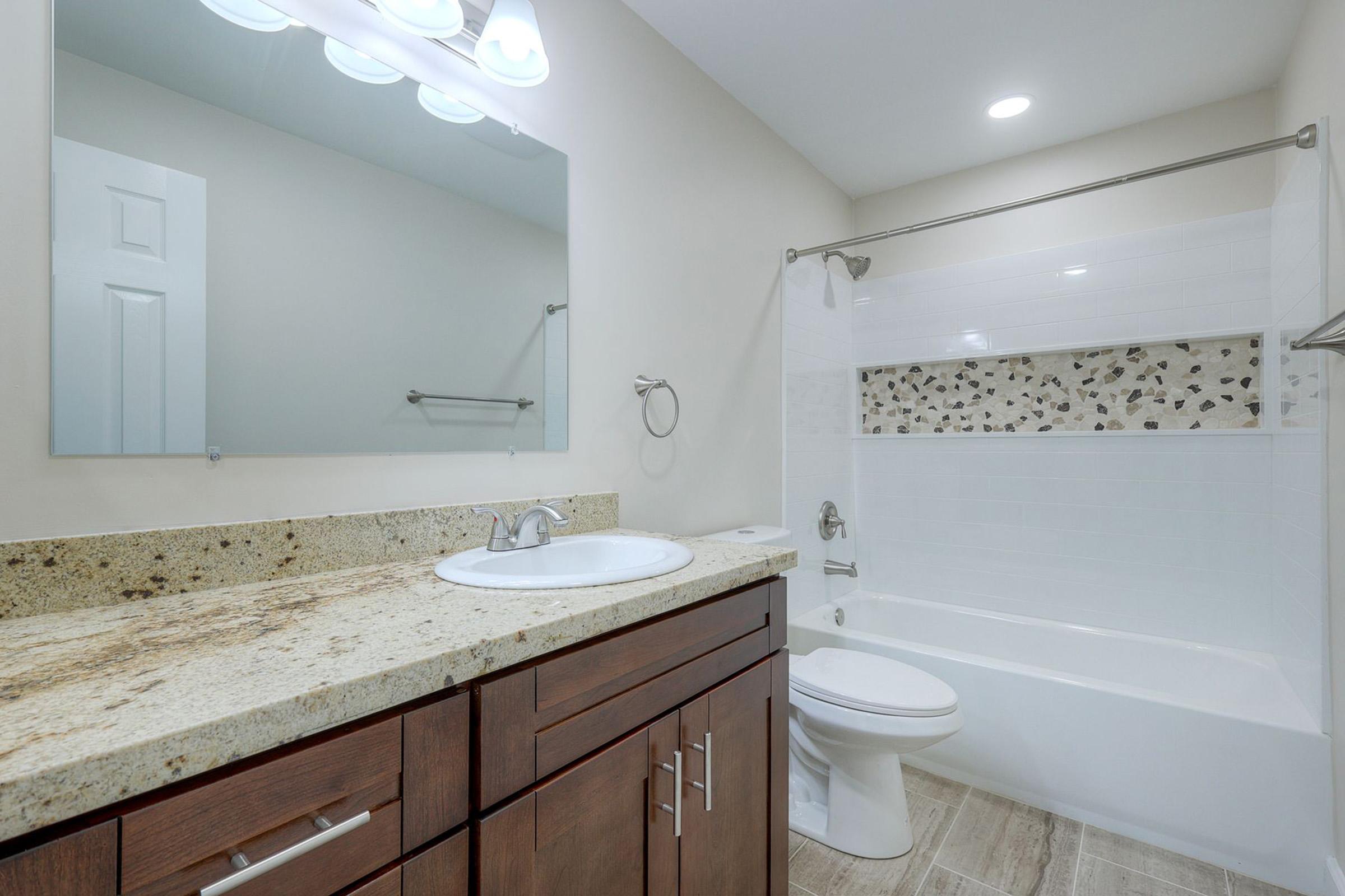 A modern bathroom featuring a granite countertop with a sink and wooden cabinetry. The adjacent tub has a white tiled surround with decorative stone accents. A large mirror hangs above the sink, and there is a shower curtain next to the tub. Soft lighting illuminates the space, creating a clean and airy atmosphere.