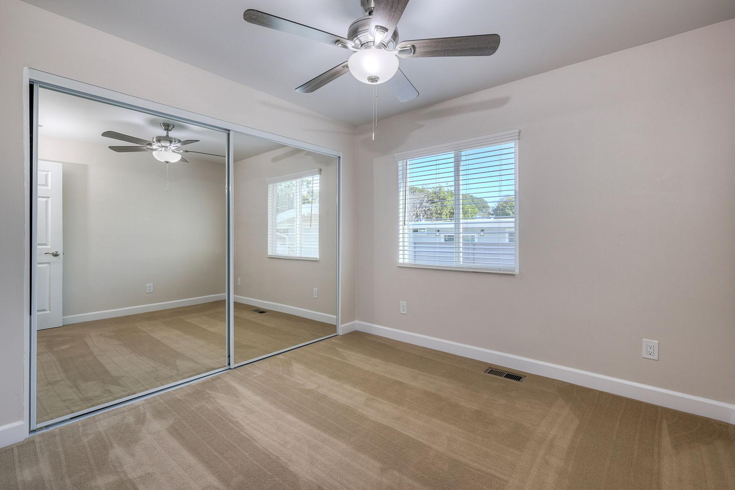 A spacious, well-lit room with a ceiling fan and large mirrored sliding closet doors. The walls are painted a light color, and there is a window providing natural light. The floor is covered with beige carpeting, creating a cozy atmosphere.