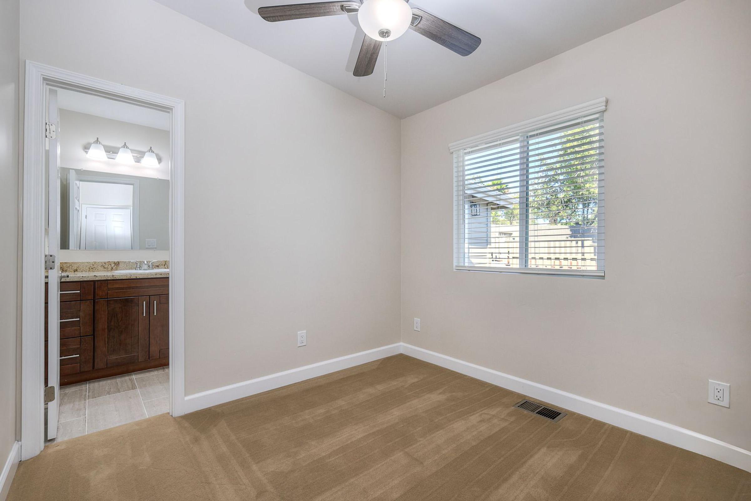 A well-lit empty room featuring beige walls and carpet. It has a ceiling fan and a window with blinds, offering a view of the outdoors. On one side, there is an open door leading to a bathroom, which includes modern fixtures and cabinetry visible in the reflection.