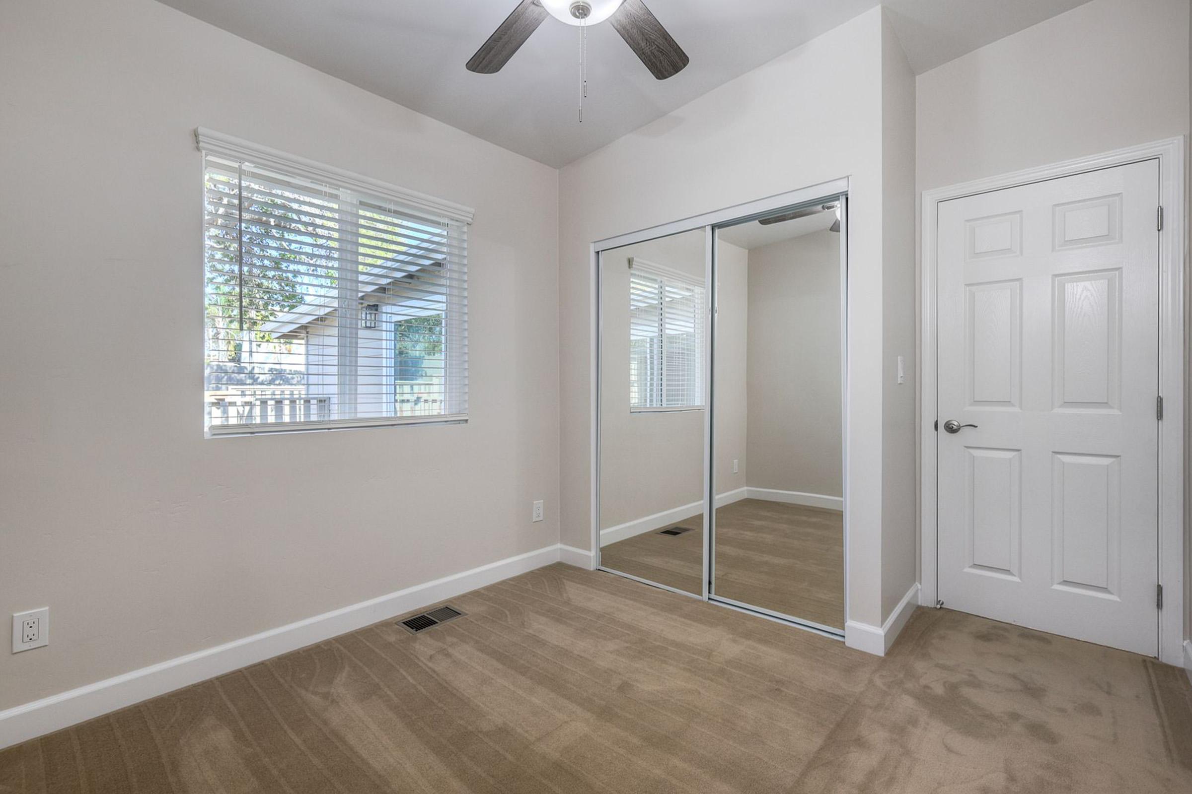 A well-lit, empty room featuring light beige walls, a window with blinds, and a ceiling fan. There is a mirrored closet door on one wall and a door leading to another space. The carpeted floor is a neutral color, contributing to a spacious and inviting atmosphere.
