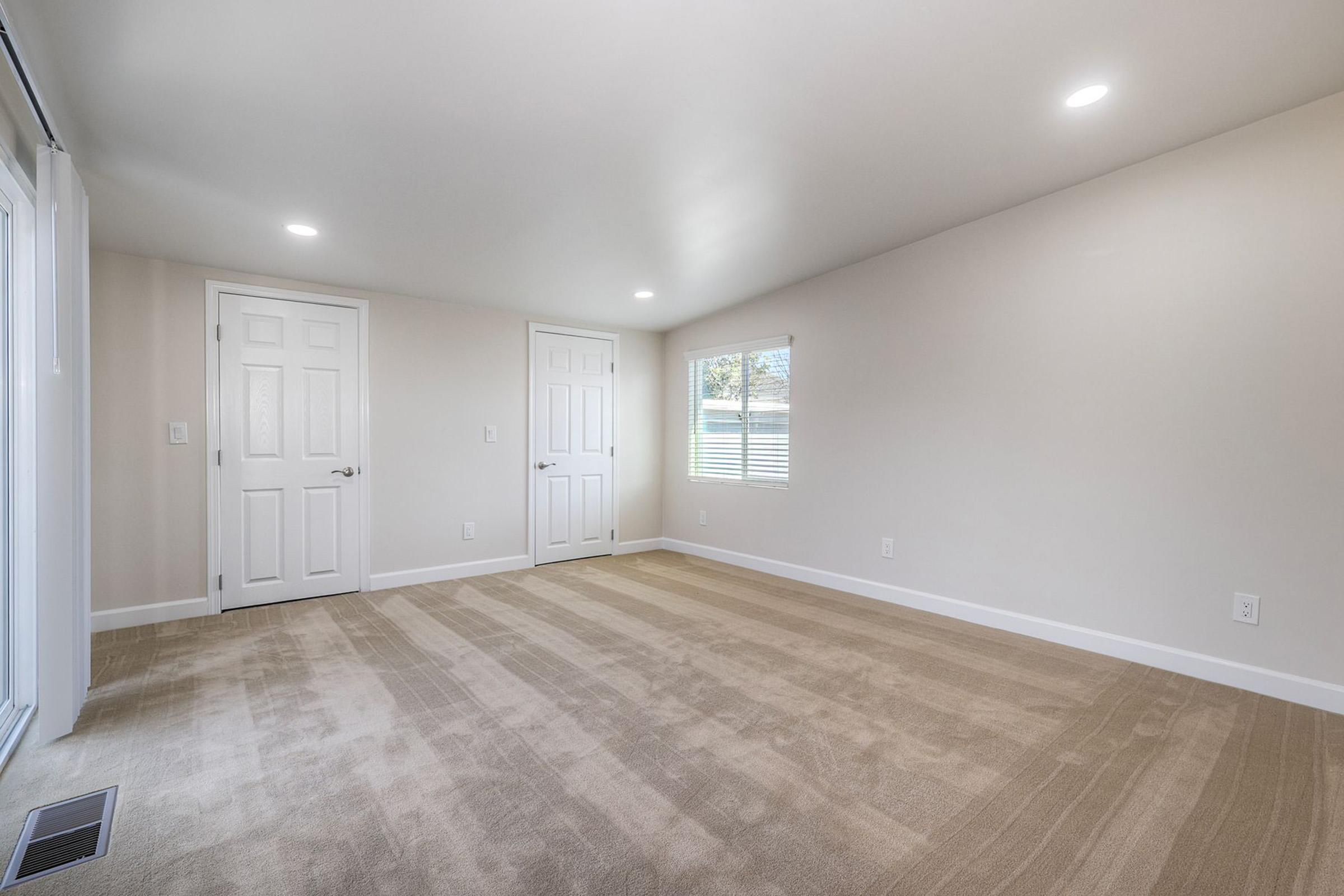 A clean, empty room with beige carpet and light-colored walls. Two large white doors are visible, one leading to an outside area. A window allows natural light to enter, casting a soft glow. The space is bright and uncluttered, ready for furniture or decoration.