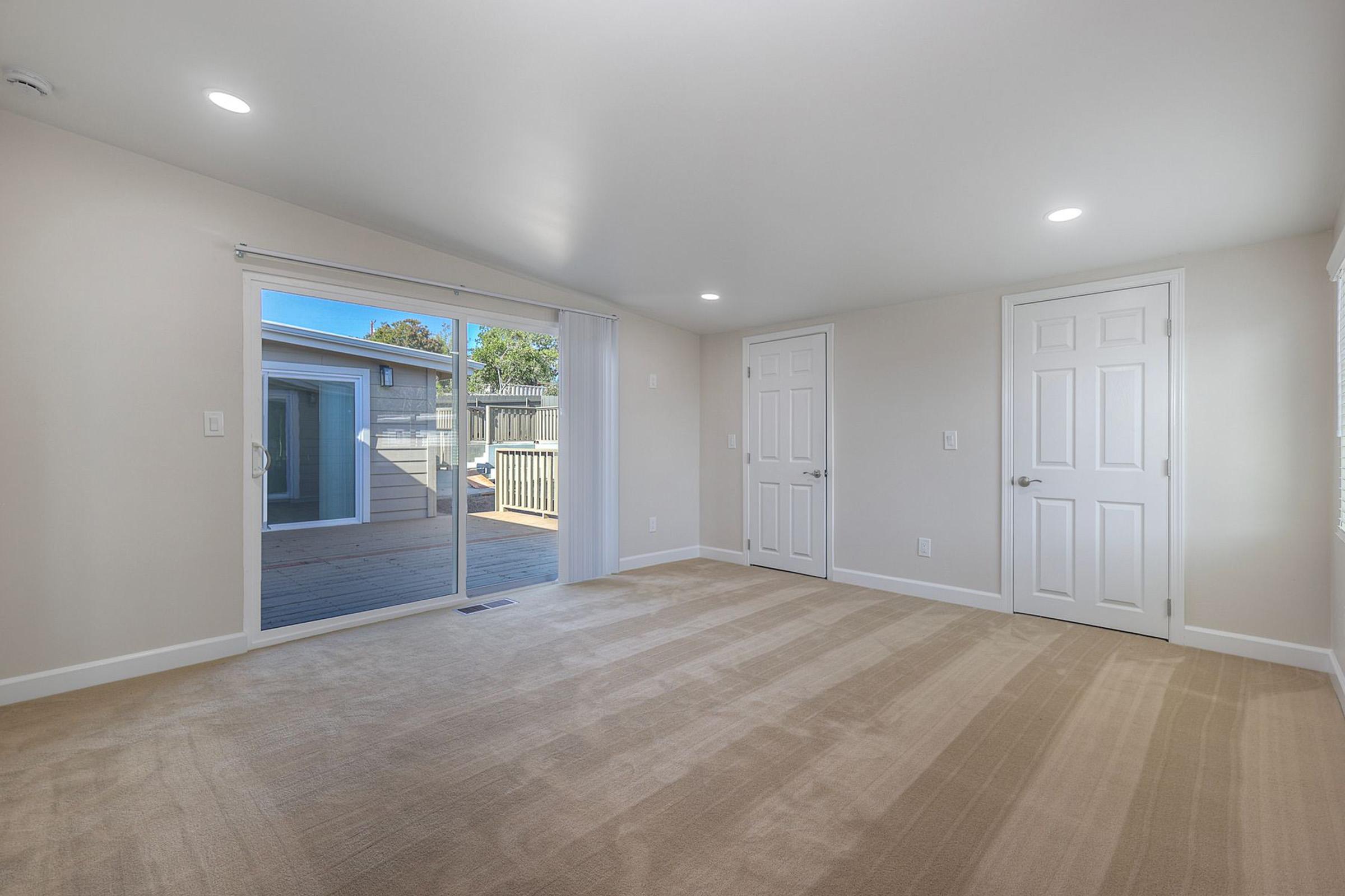 A bright, empty room featuring beige carpet, white walls, and two closed doors. A sliding glass door leads to an outdoor deck area. The room is well-lit with natural light streaming in, offering a spacious and clean environment.