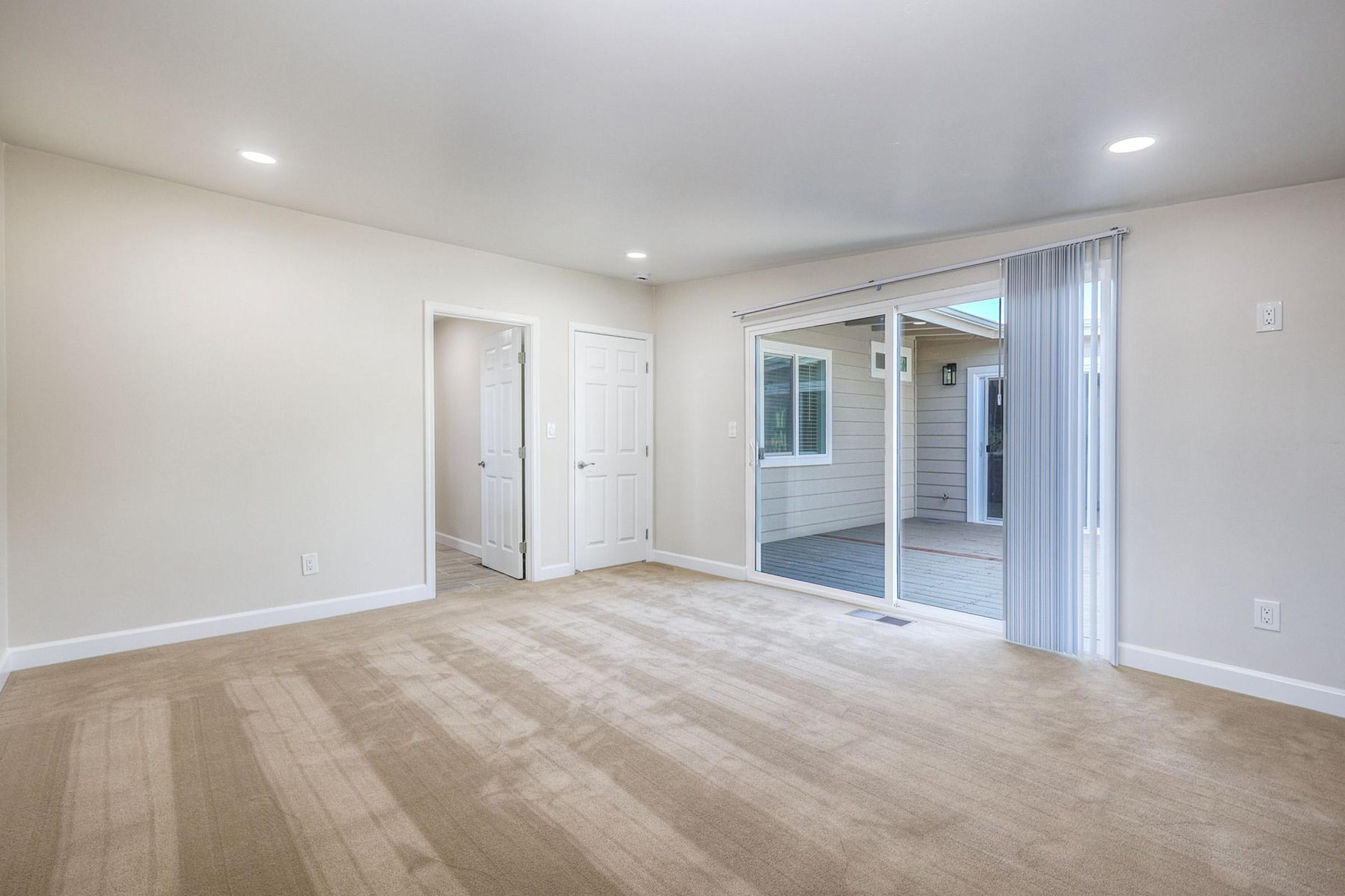 A bright, empty living room with beige carpet and light-colored walls. There is a sliding glass door leading to an outdoor patio, and a doorway visible on the left leading to another room. The space is well-lit with recessed ceiling lights.