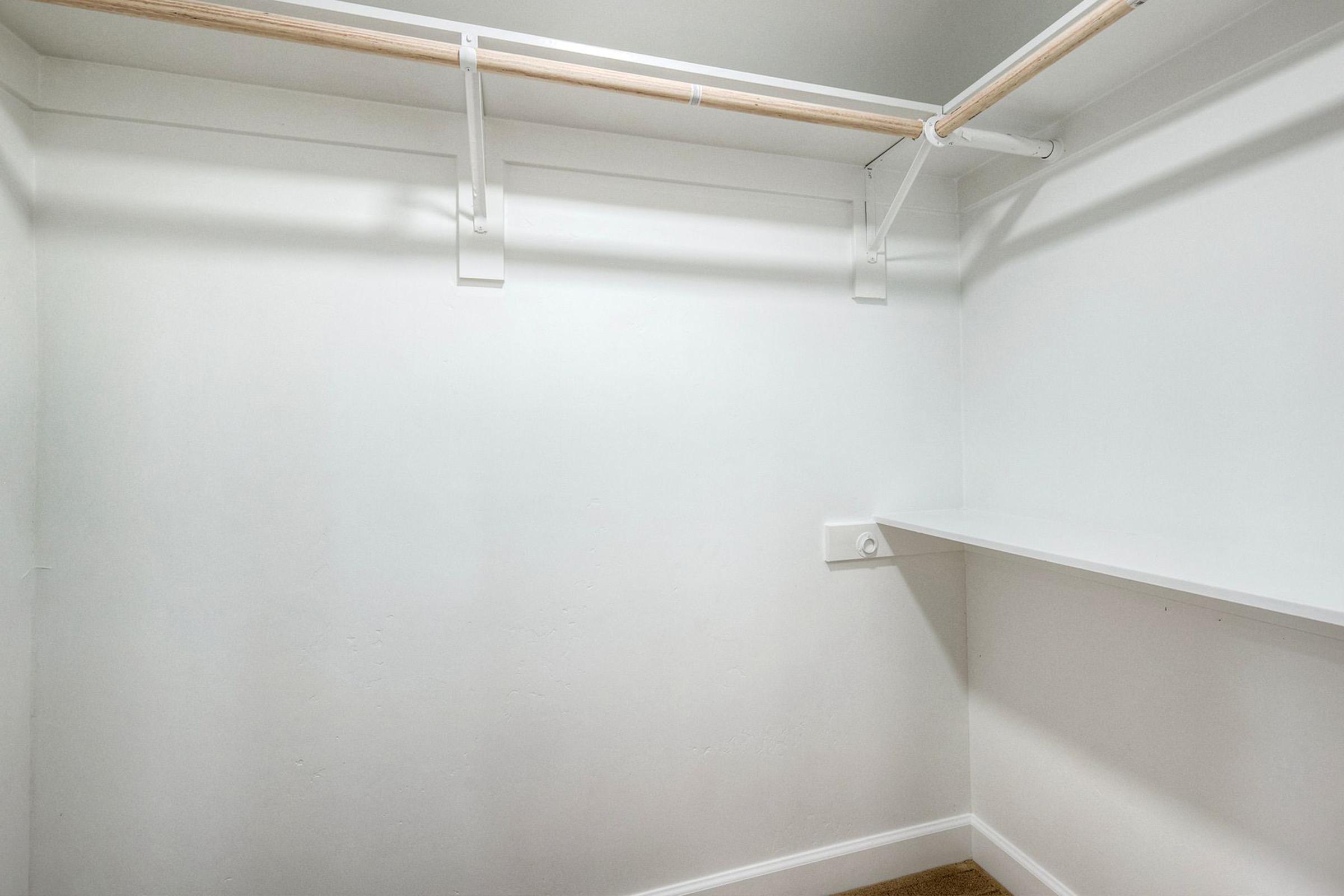 Empty white closet featuring a wooden rod for hanging clothes and a built-in shelf along one side. The walls are plain and unadorned, emphasizing the spaciousness of the area.