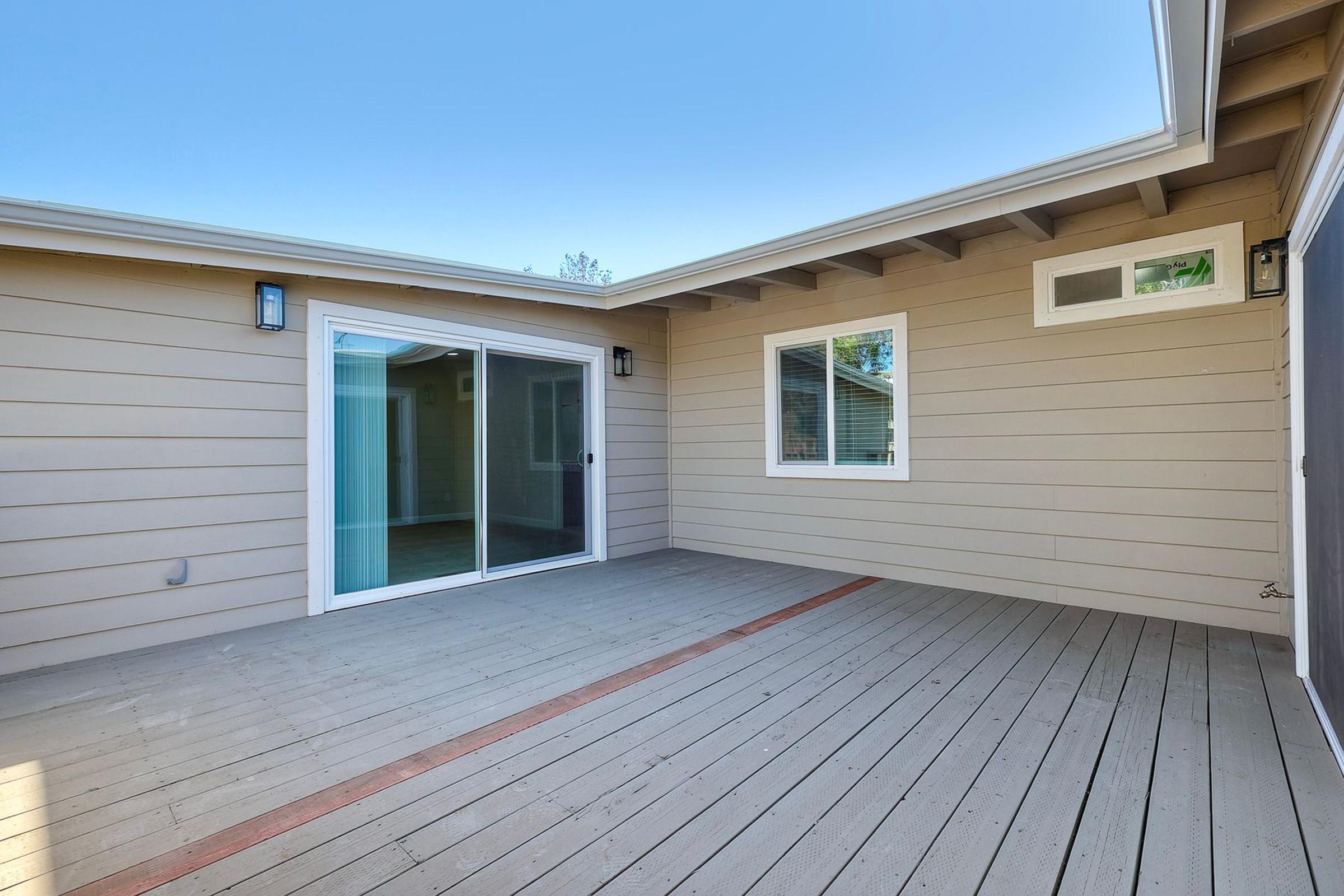A spacious, modern outdoor patio area featuring wooden flooring, sliding glass doors, a few windows, and light-colored walls, set against a clear blue sky. The area appears well-lit and inviting, ideal for relaxation or social gatherings.