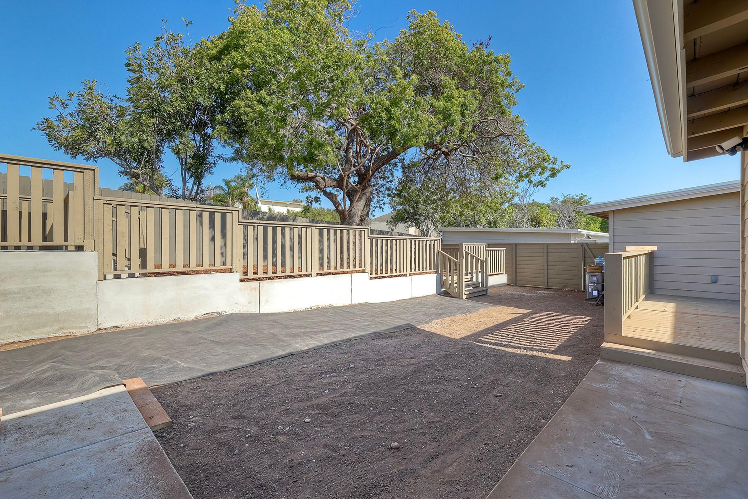 A spacious outdoor area featuring a bare dirt patch, surrounded by wooden railings and a large tree. Sunlight casts shadows on the ground, while a building is partially visible on the right. The area is ready for landscaping or further development.