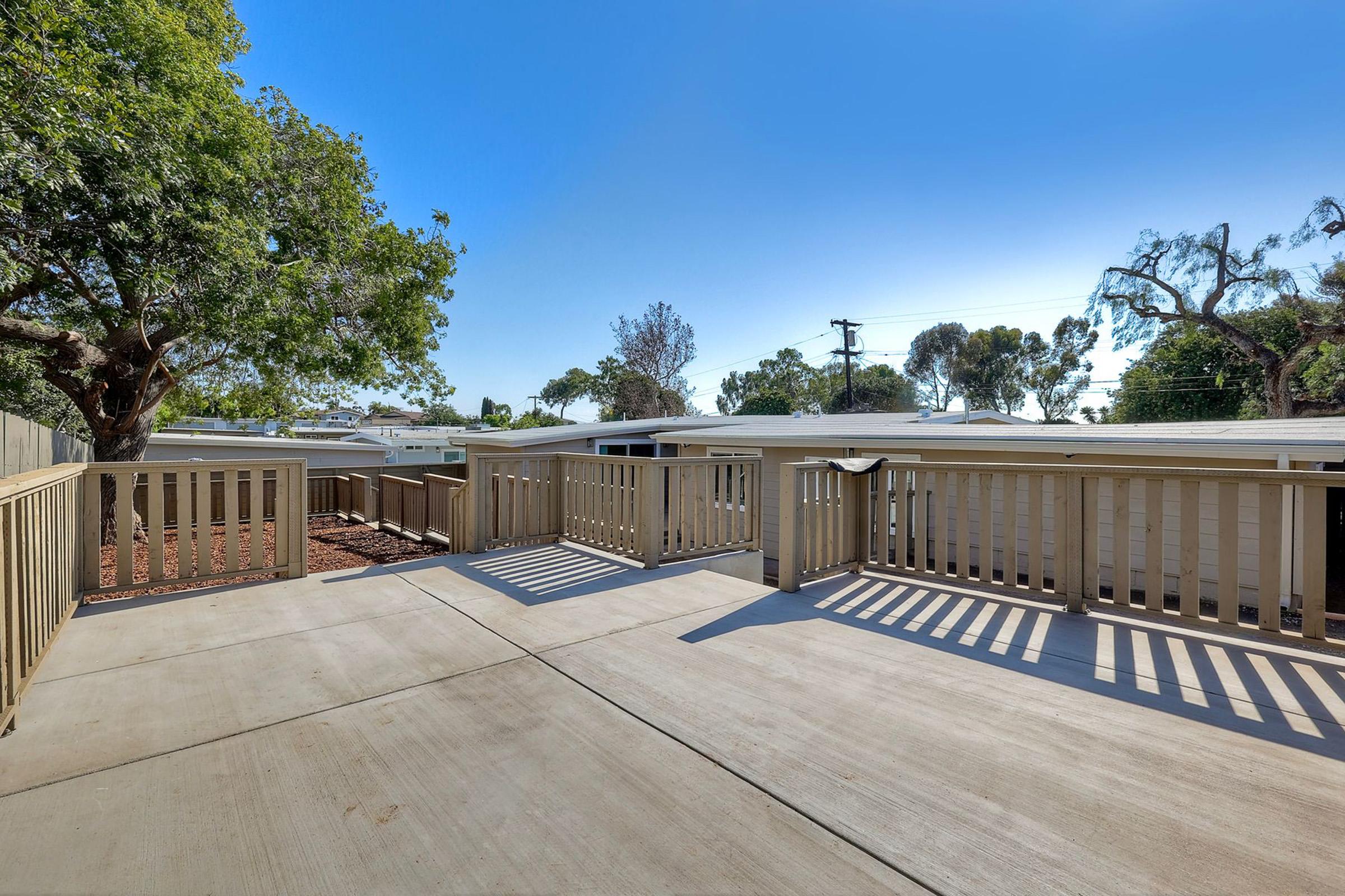 A spacious outdoor deck with wooden railings, surrounded by trees and a clear blue sky. The concrete surface is well-maintained, leading to a quiet residential area with houses in the background. Sunlight casts shadows on the deck, creating a welcoming atmosphere.