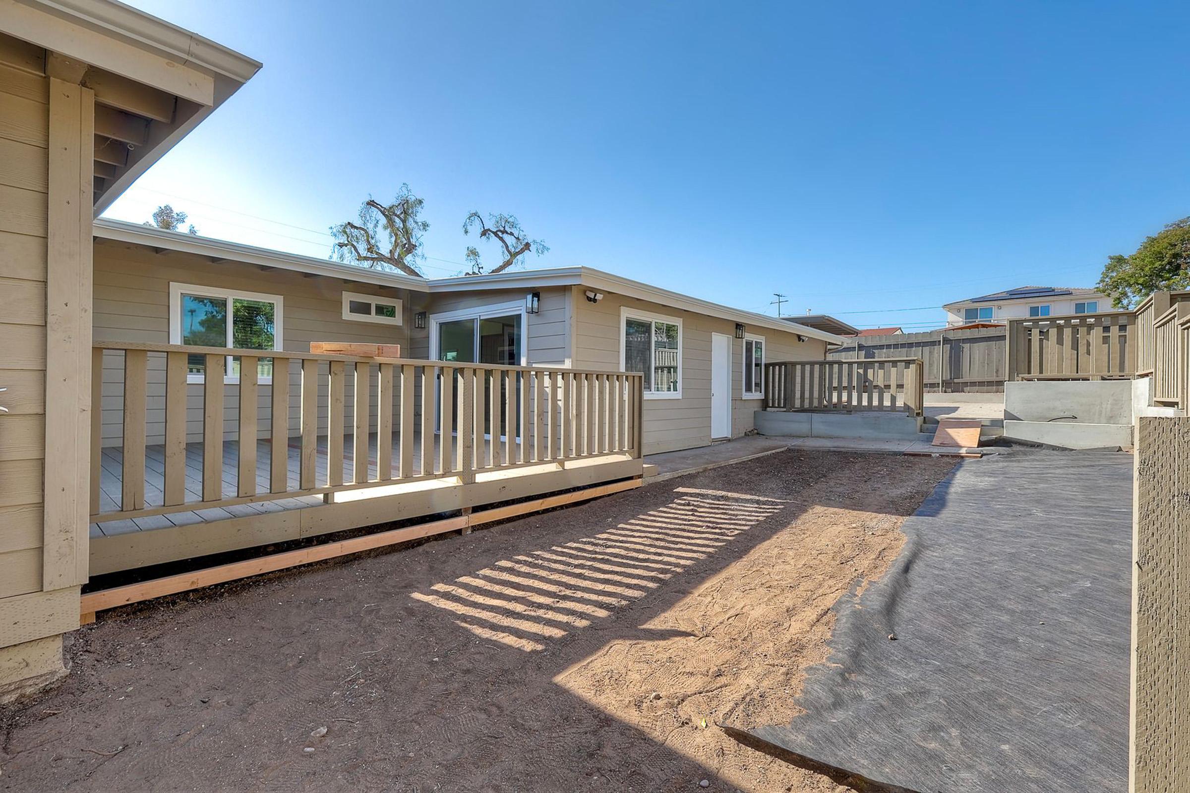 A residential backyard featuring a paved area and wooden deck, with newly constructed beige exterior walls and several windows. The space is partially covered with black landscaping fabric, and a fence is visible in the background. The scene is set under a clear blue sky.