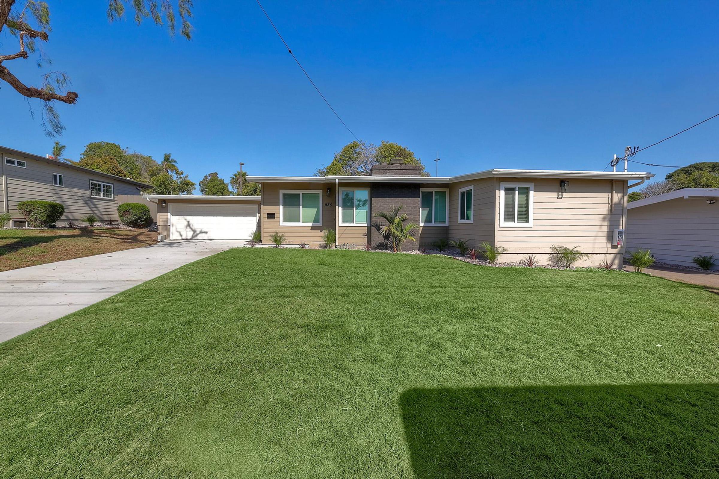 A modern single-story home with a well-manicured lawn, featuring large front windows, a stone accent wall, and a paved driveway. The house is surrounded by trees and shrubs, and there is a carport on the left side. The clear blue sky enhances the welcoming appearance of the property.