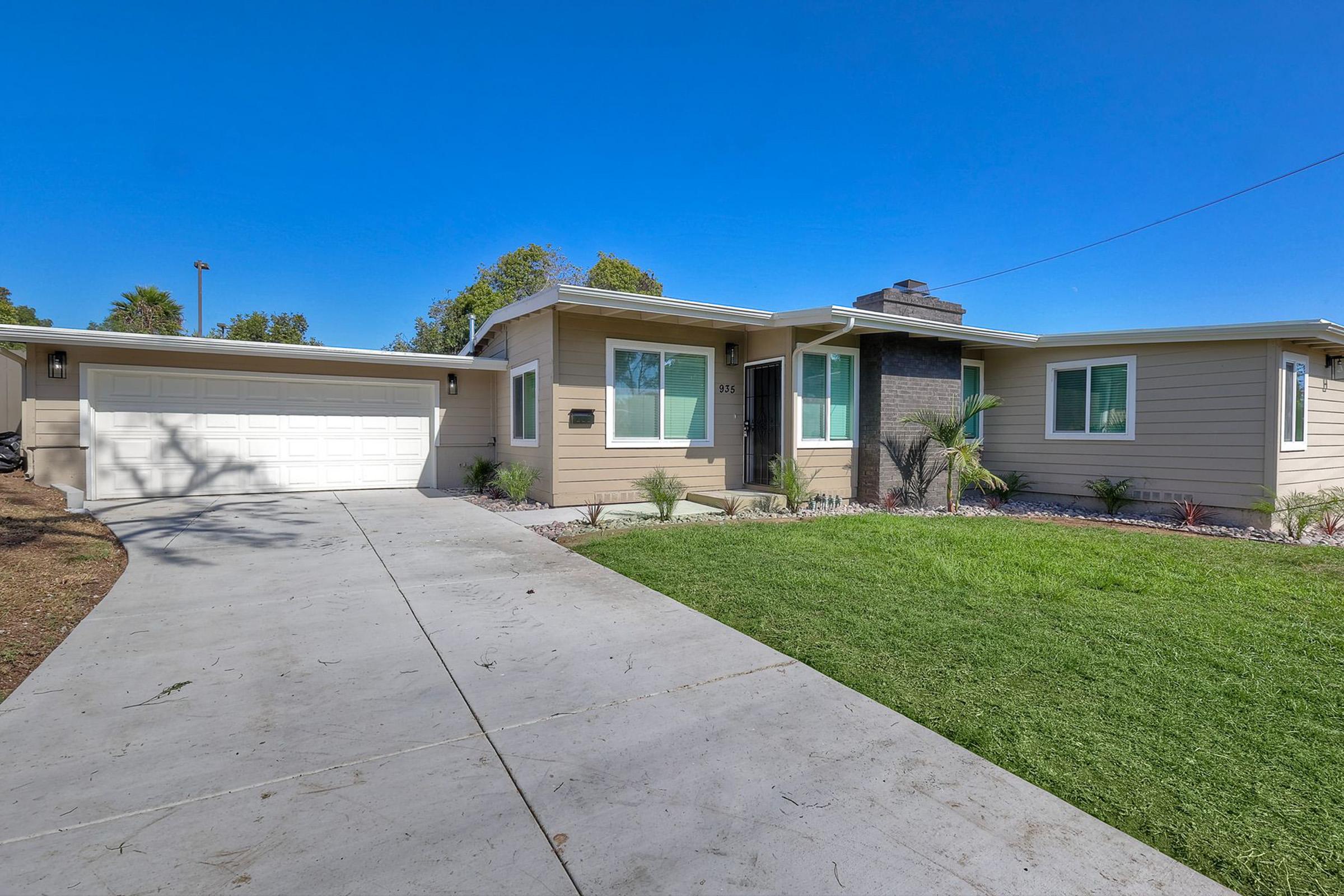 A modern single-story house with a light beige exterior, large windows, and a charcoal stone accent wall. The home features a neatly landscaped front yard with grass and plants. A paved driveway leads to a two-car garage on the left side. Bright blue sky overhead adds to the cheerful appearance of the property.